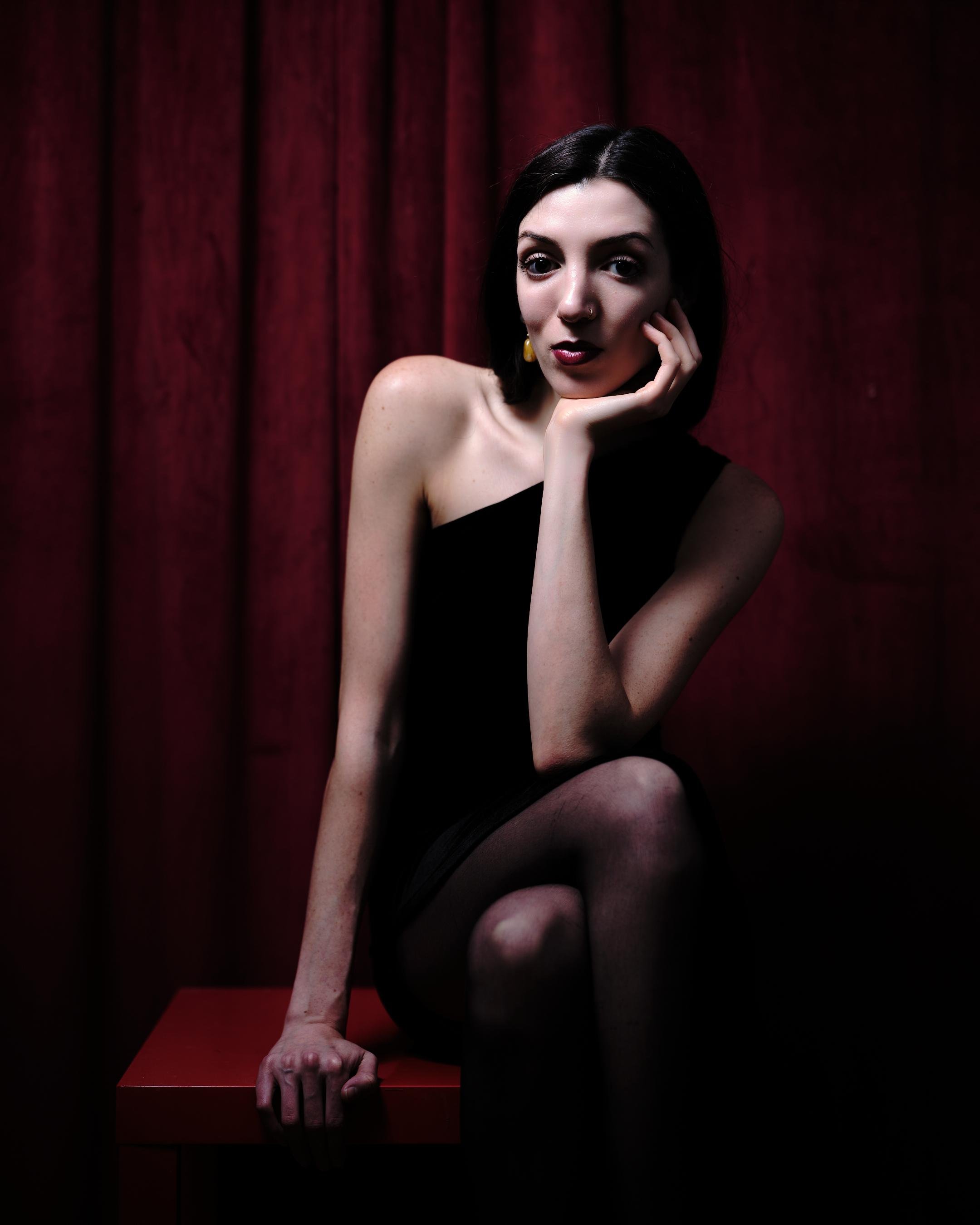 Woman in a black one-shoulder dress and dark tights sits on a red bench, resting her chin on her hand, against a burgundy curtain backdrop in low-key lighting.