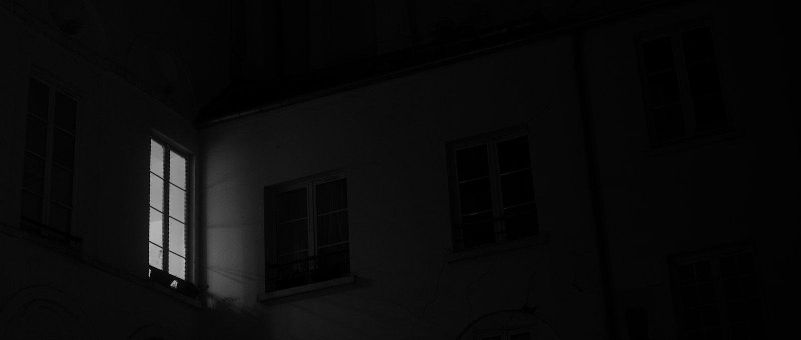 Photo horizontale format widescreen, noir et blanc. Sur la gauche, une unique fenëtre dont de la lumière émane. On distingue d’autre fenêtres dans l’obscurité, sombres