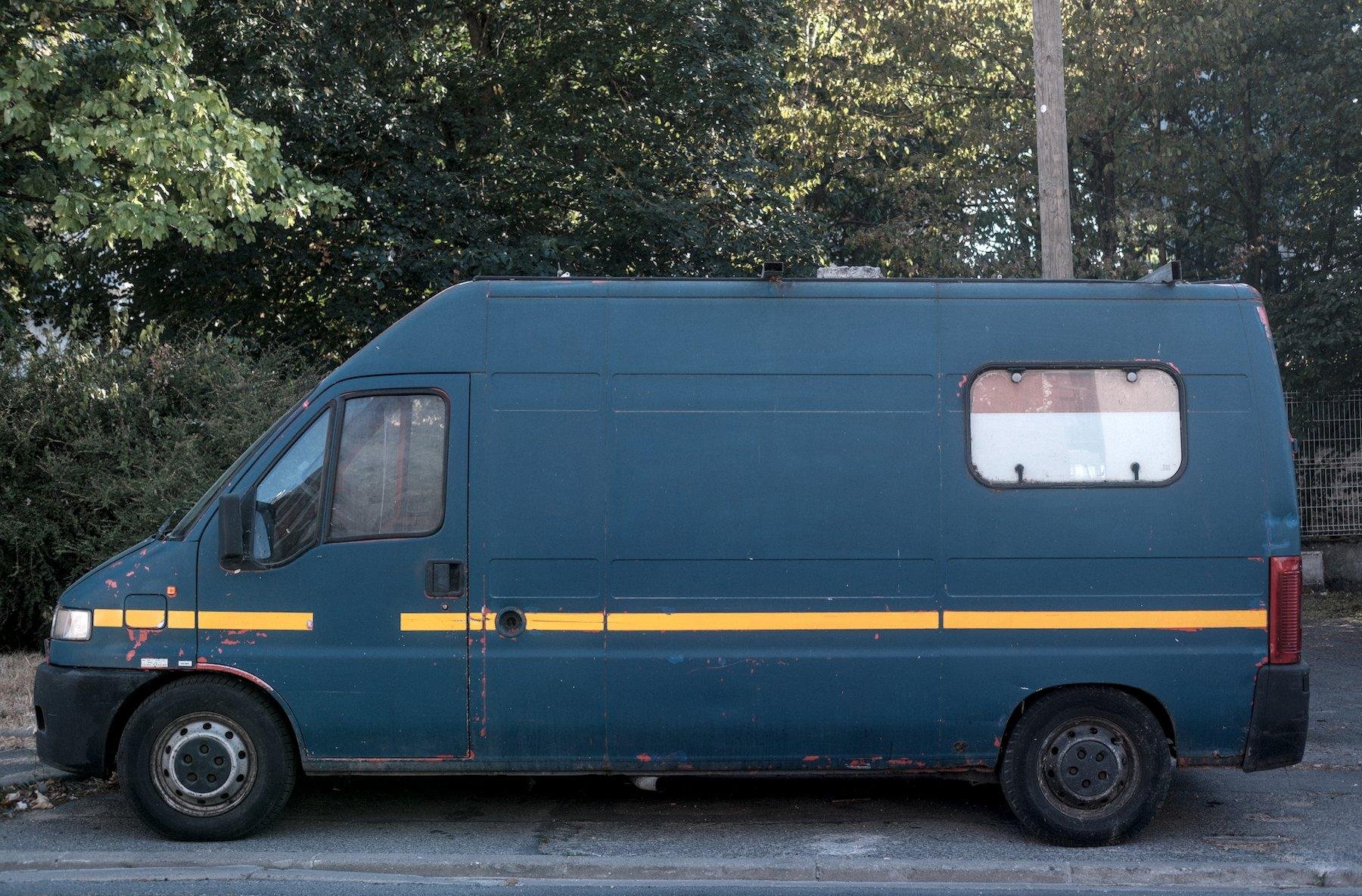 Photo horizontale. Une camionnette garée dans la rue, de couleur bleu/vert avec une bande jaune horizontale juste en dessous de la poignée de portière, sur toute la longueur du véhicule