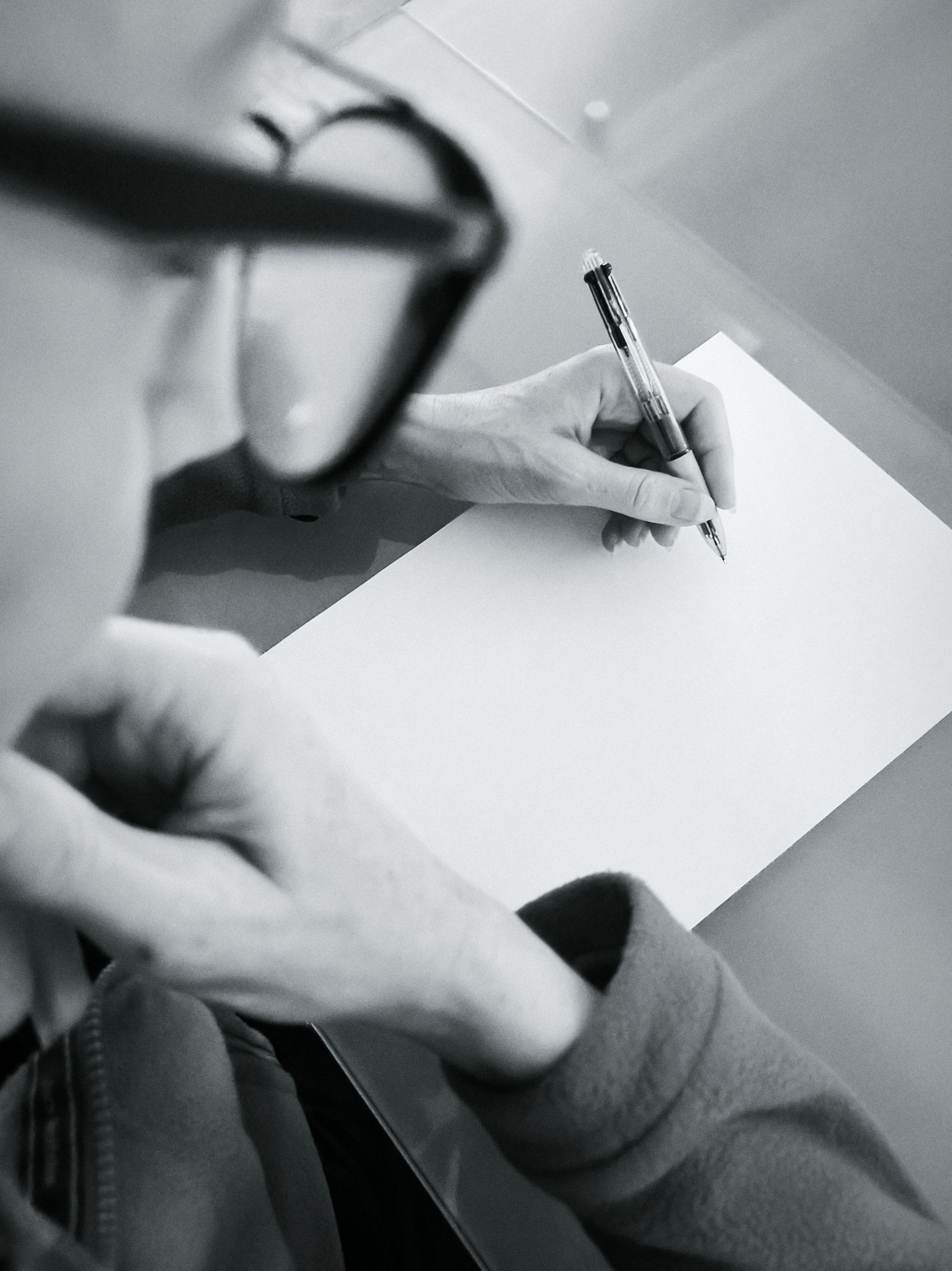 Over-the-shoulder black-and-white close-up of a person in glasses holding a pen above a blank sheet of paper on a desk. 
### 
Schwarzweiß-Nahaufnahme über die Schulter einer Person mit Brille, die einen Stift über einem leeren Blatt Papier auf einem Schreibtisch hält. 