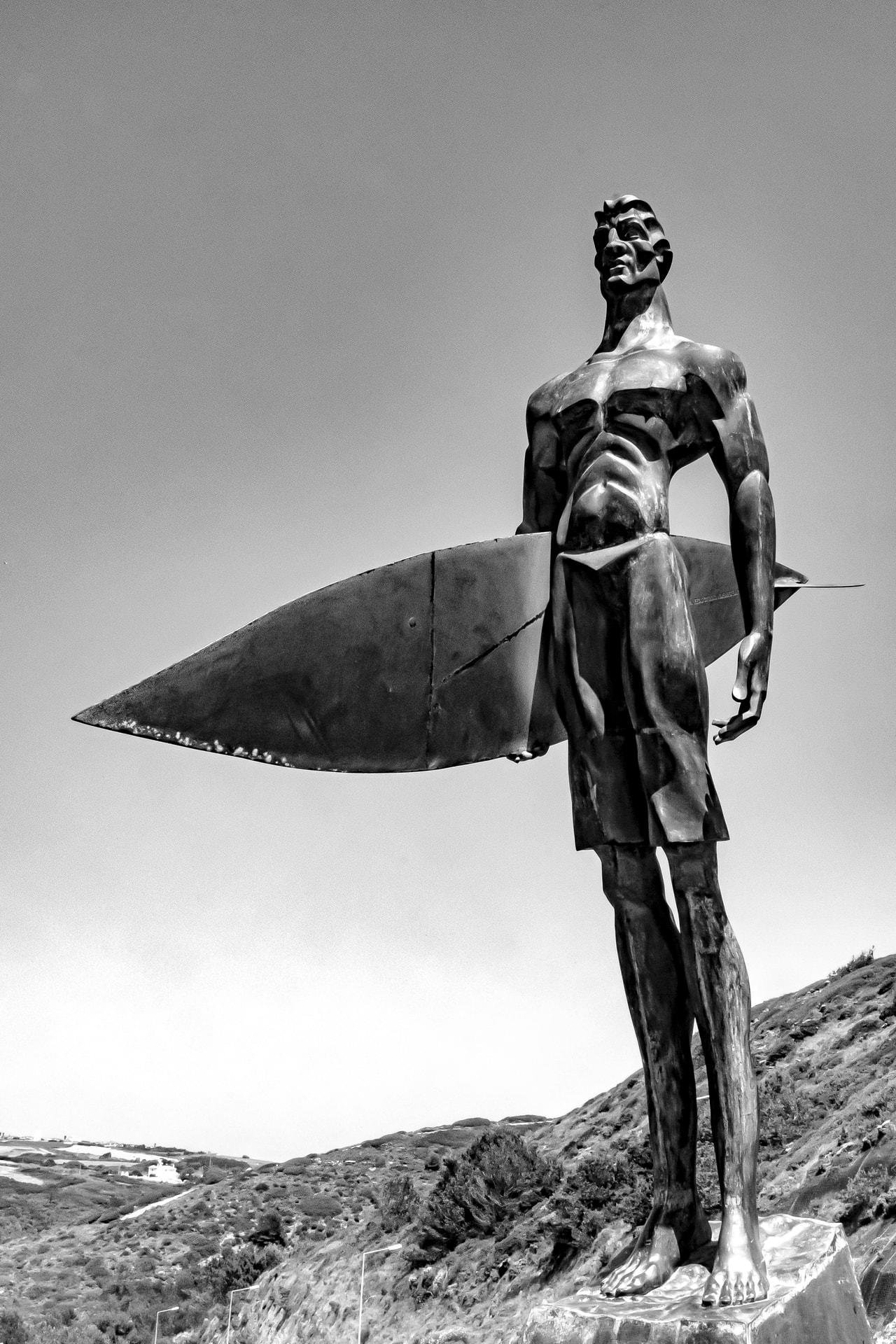 Black-and-white low-angle view of a tall metal statue of a shirtless surfer holding a surfboard under one arm, standing on a pedestal with hills and coastline in the background and open sky above.