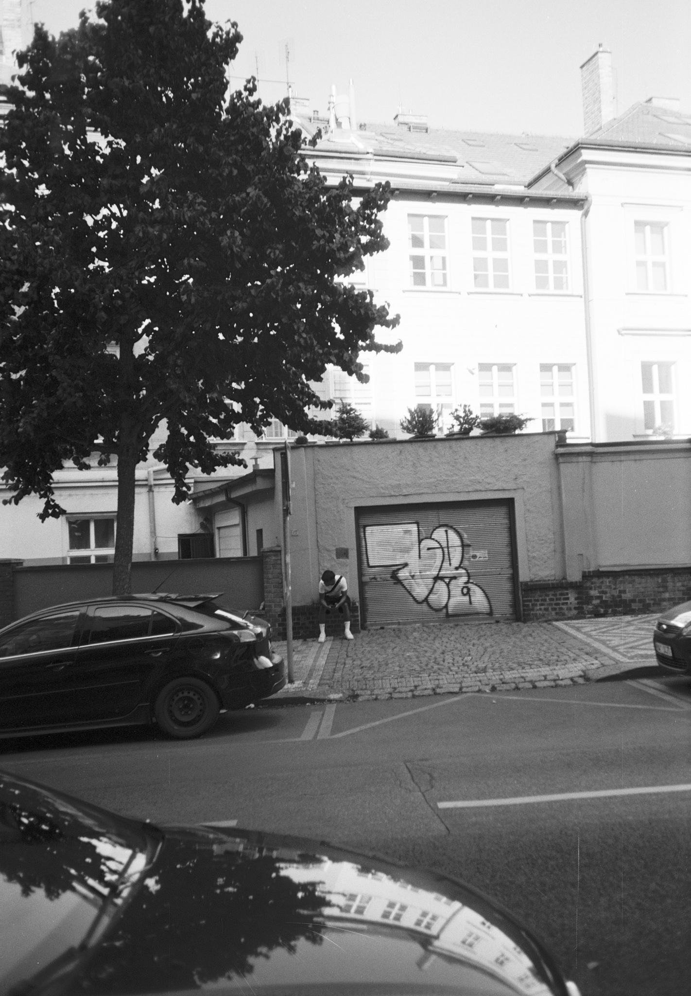 Black-and-white street scene with parked cars along a curb, a large leafy tree on the left, and a low garage building with a roll-up door covered in graffiti; a person sits on the curb near a pole in front of the garage, with a multi-story residential building and rows of windows in the background.