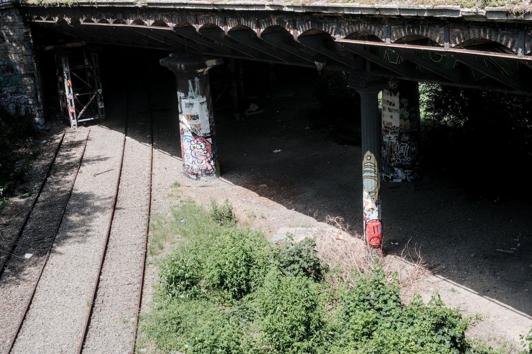 Photo horizontale. L’image est partagée en deux sur l’axe NW/SE. En bas à gauche, des 2 voie de rails et des buissons en plein soleil, en haut à droite, un pont et ses pilier tagués met le sol dans l’ombre