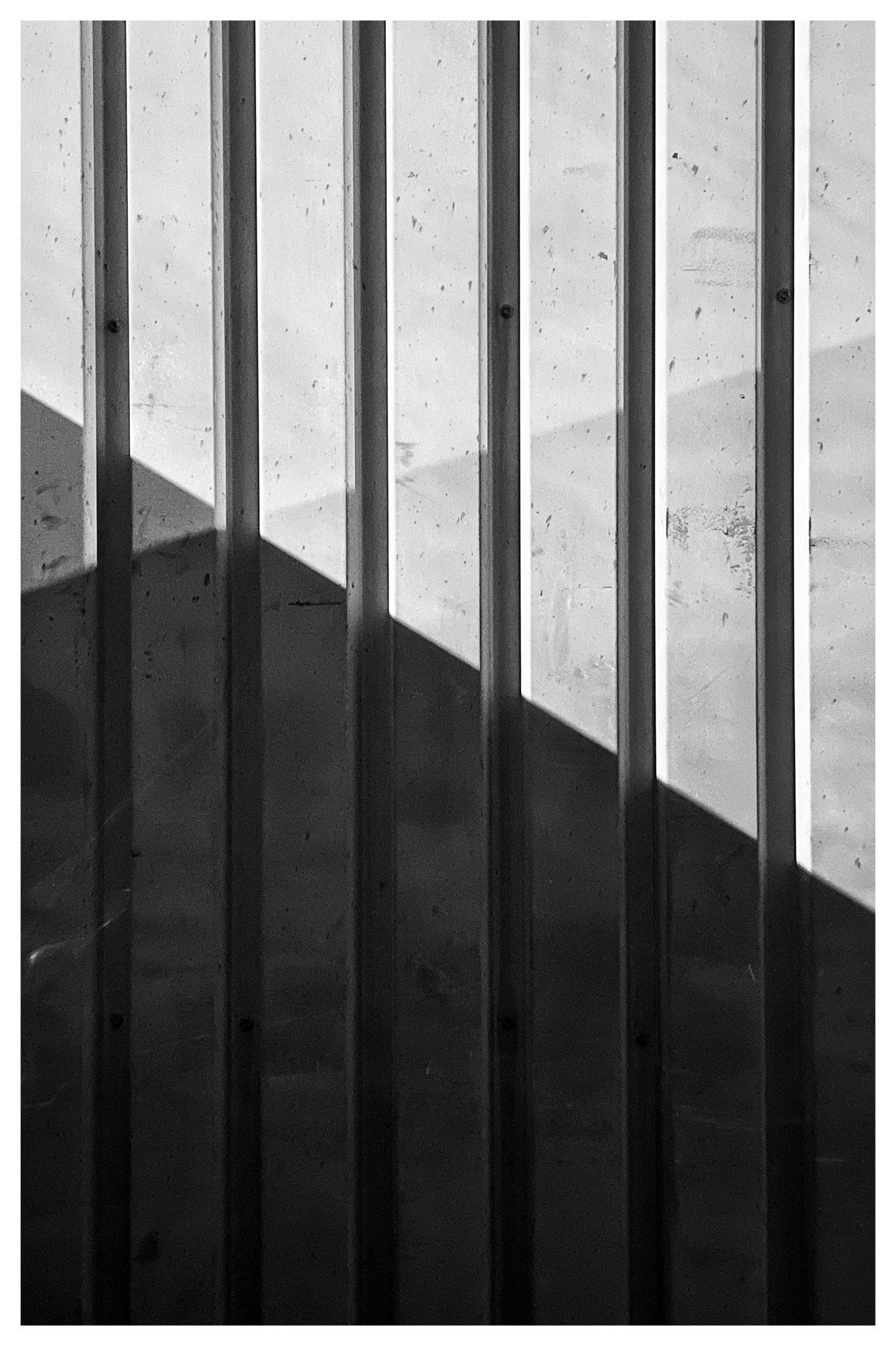 Vertical metal studs against a light wall, with a strong diagonal shadow cutting across the lower half of the frame.