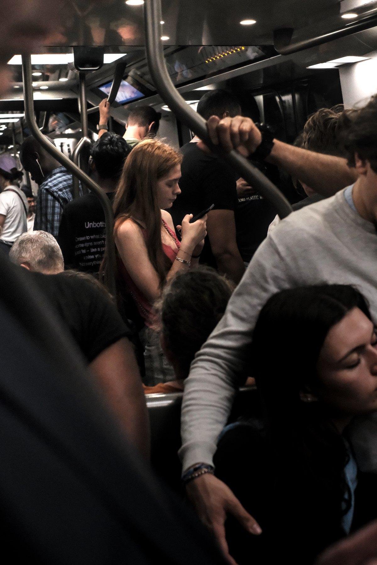 Photo verticale. Dans une rame de métro, debout au milieu de gens en noir ou en blanc, une jeunne femme rousse au cheveux long lâchés en débardeur rose regarde son téléphone