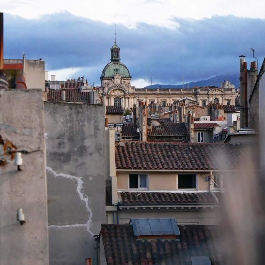 L'image représente une vue d'un paysage urbain avec un accent sur les toits et le bâtiment architectural au loin, de la préfecture des Bouches-du-Rhône. Les toits sont recouverts de tuiles rouges traditionnelles et le bâtiment à l'arrière-plan est coiffé d'un dôme et orné de détails. Le ciel est nuageux, ce qui ajoute de l'ambiance à la scène urbaine.