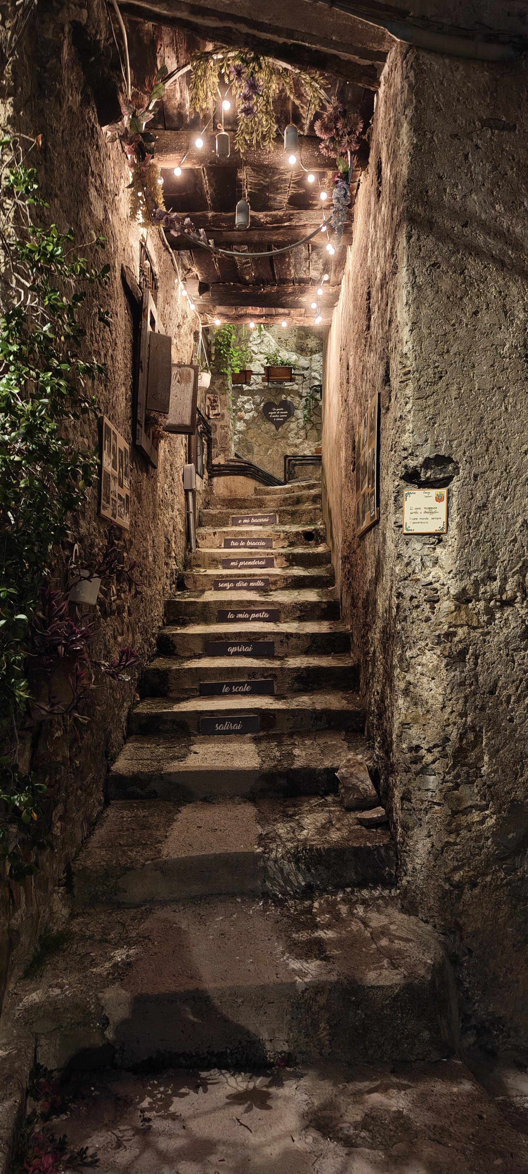 Stretto vicolo notturno con scale di pietra, muri invecchiati, edera sul lato sinistro, luci decorative calde visibili in alto nel sottofondo