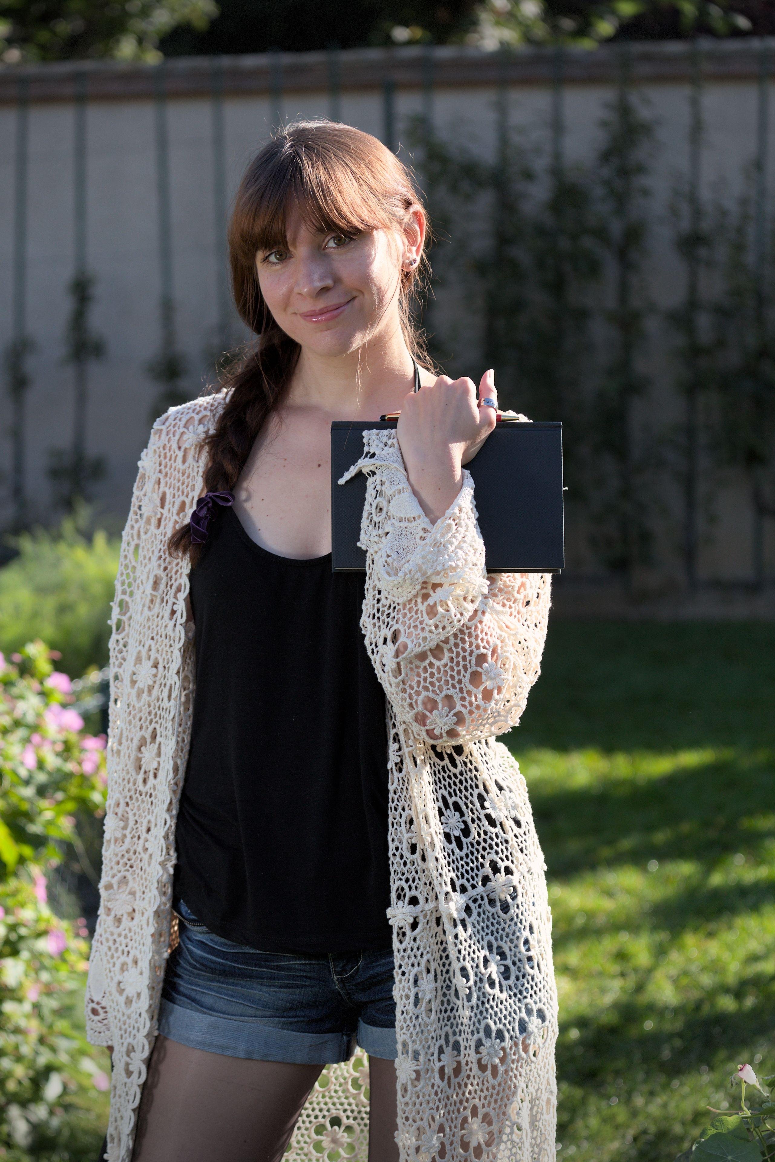 une fille châtain, debout, cheveux coiffés en une natte qui arrive sous les épaules, regardant la caméra et tenant fermement un carnet de notes. Elle est habillé d’un débardeur noir, un mini-short en jean, des collants, et un long gilet blanc en mailles très larges formant des fleurs