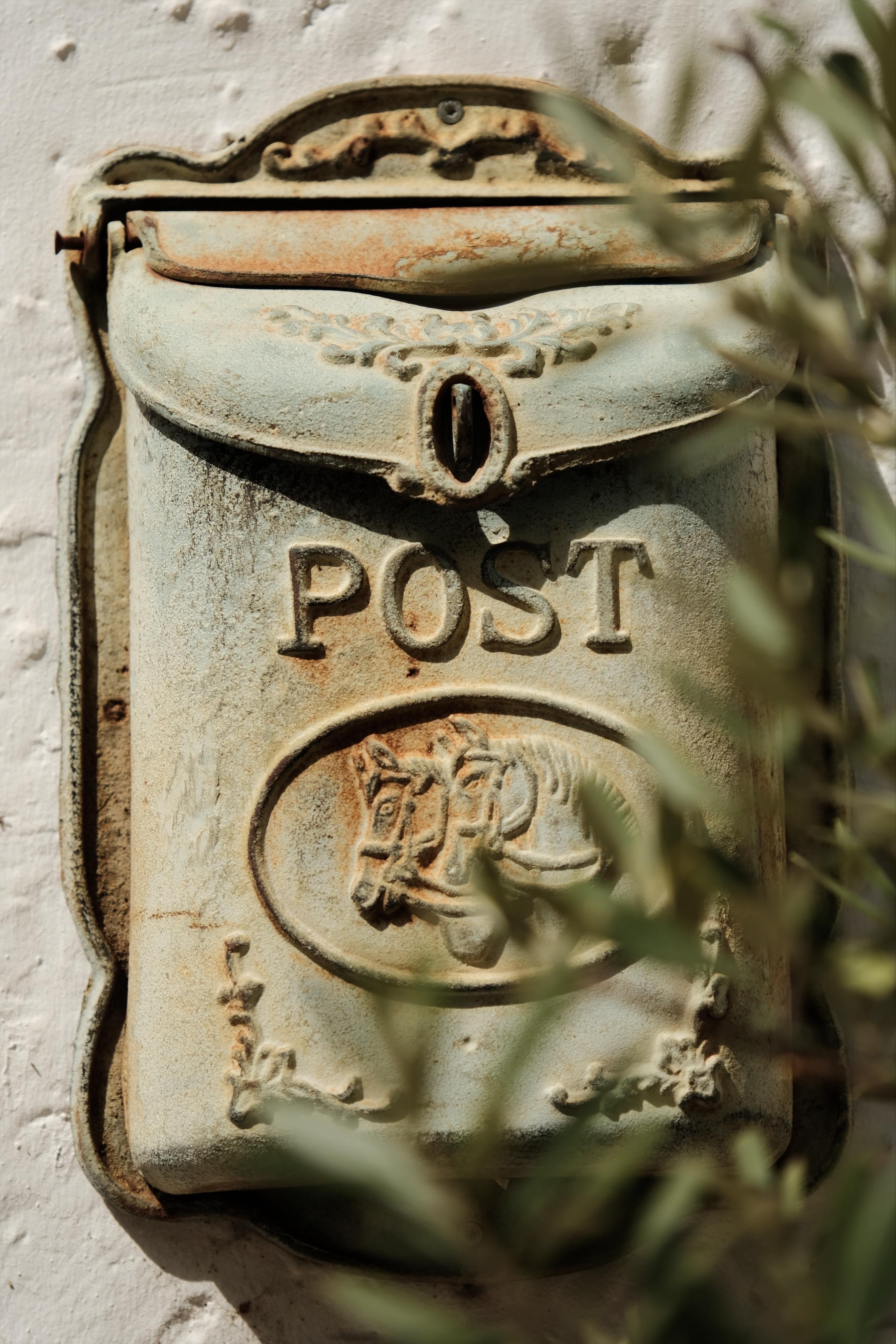 Close-up of a weathered metal wall-mounted mailbox with rust along the edges, raised “POST” lettering and a horse-head emblem, partially obscured by out-of-focus olive leaves on the right.