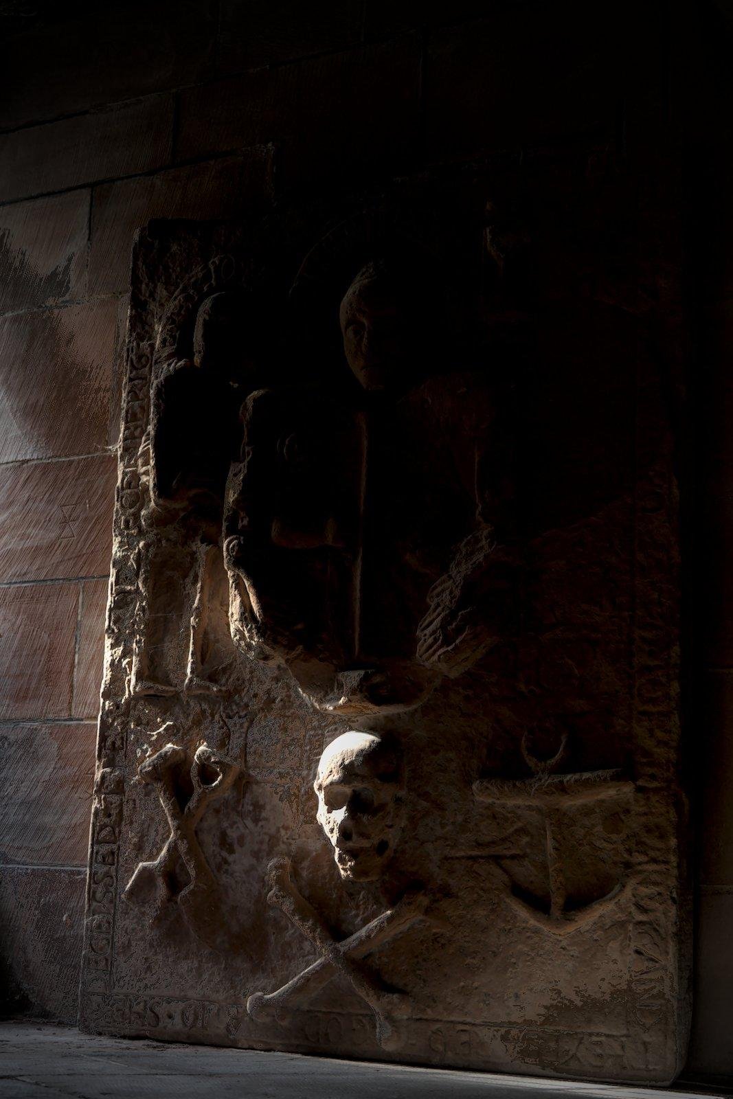 Photo verticale. Un vieux couvercle en pierre de cerceuil appuyée contre un mur, arborant des sculptures de têtes de mort, d’ancre et autre symboles marins, éclairé par un rayon de lumière arrivant de la gauche donnant un aspect dramatique à l’ensemble