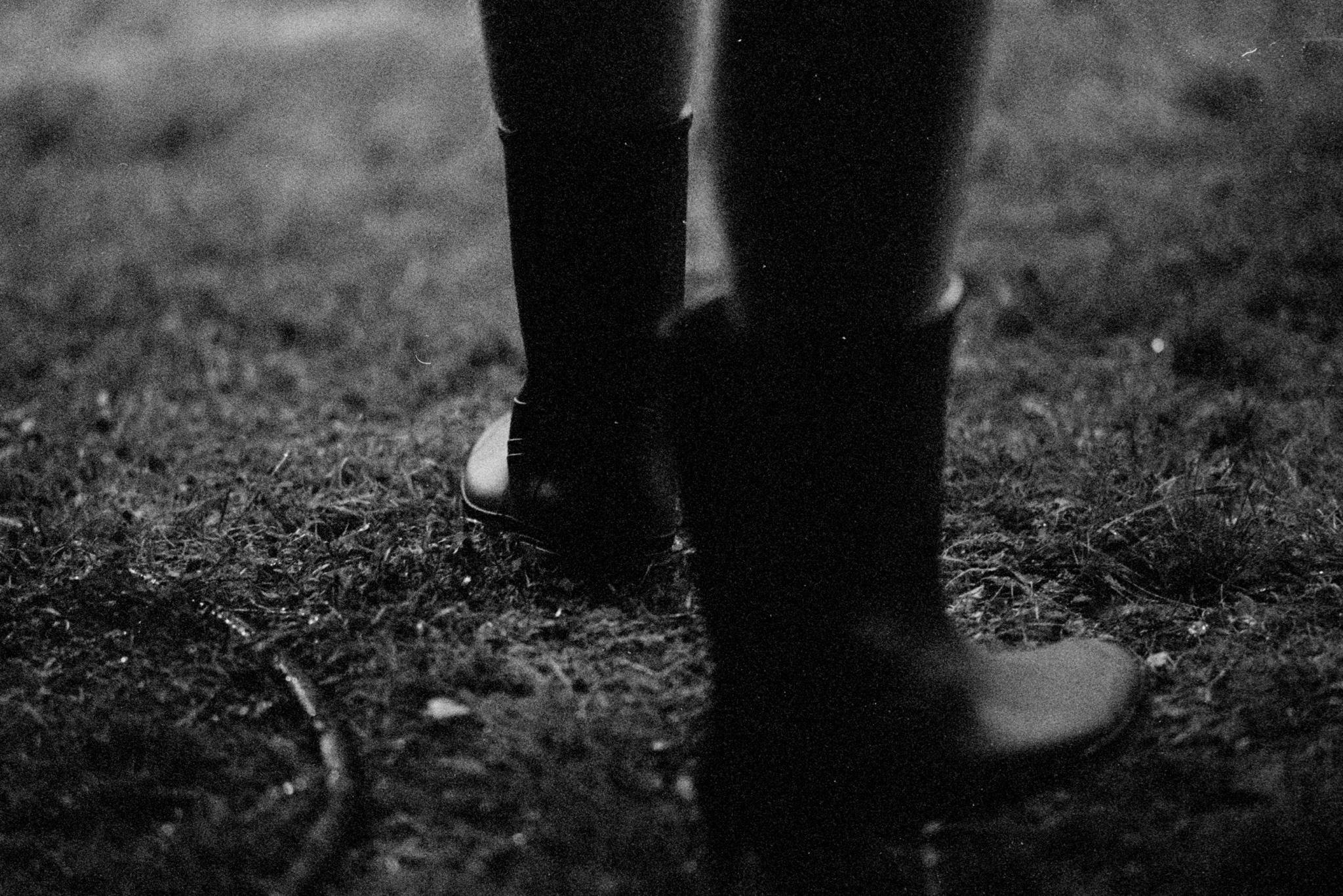Close-up black-and-white photo of a person’s lower legs in tall rubber boots standing on grass, with one boot in the foreground and the other slightly behind.