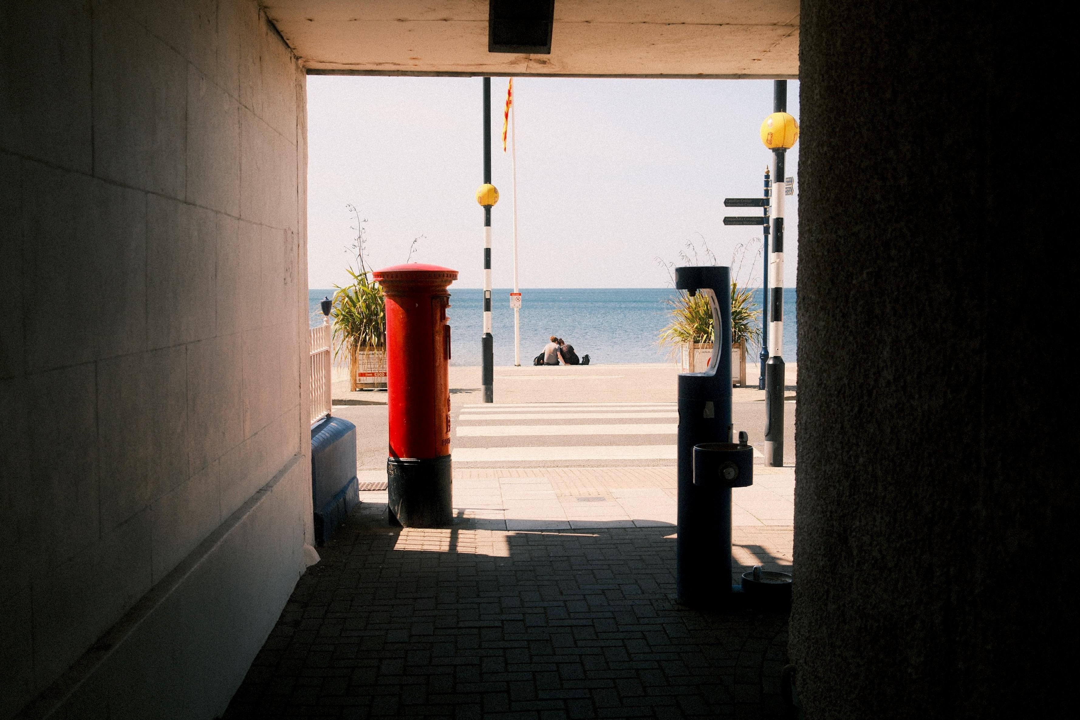Shaded passageway framing a seaside view, with a bright red cylindrical post box in the foreground, a crosswalk and street poles ahead, and two people sitting near the water on the horizon.
