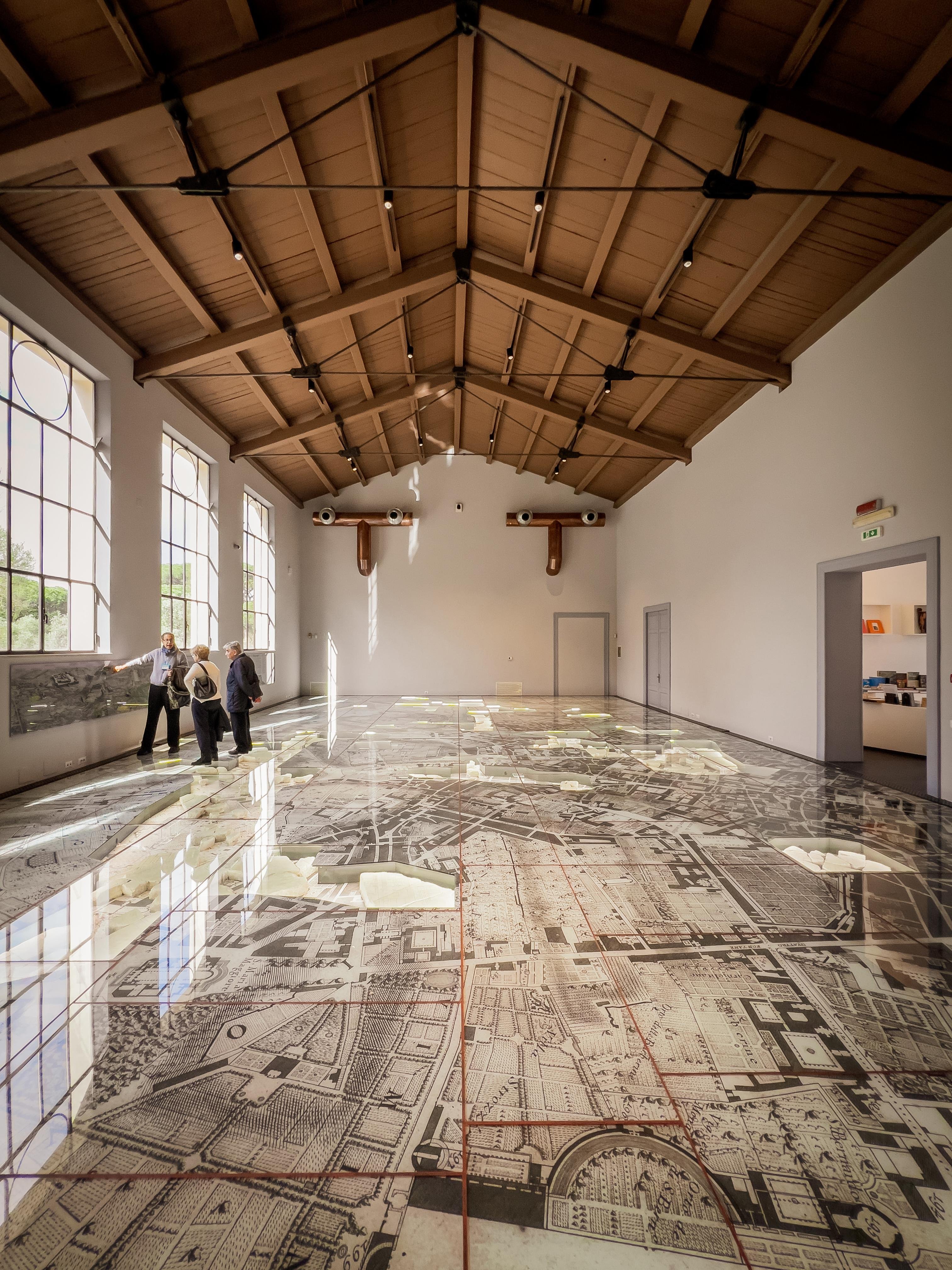 Grande stanza con un alto soffitto in legno e grandi finestre su un lato. Il pavimento ha un design dettagliato di una mappa, probabilmente rappresentando la disposizione di una città. Ci sono diverse persone in piedi che osservano la mappa sul pavimento. La stanza sembra far parte di un museo o uno spazio espositivo.