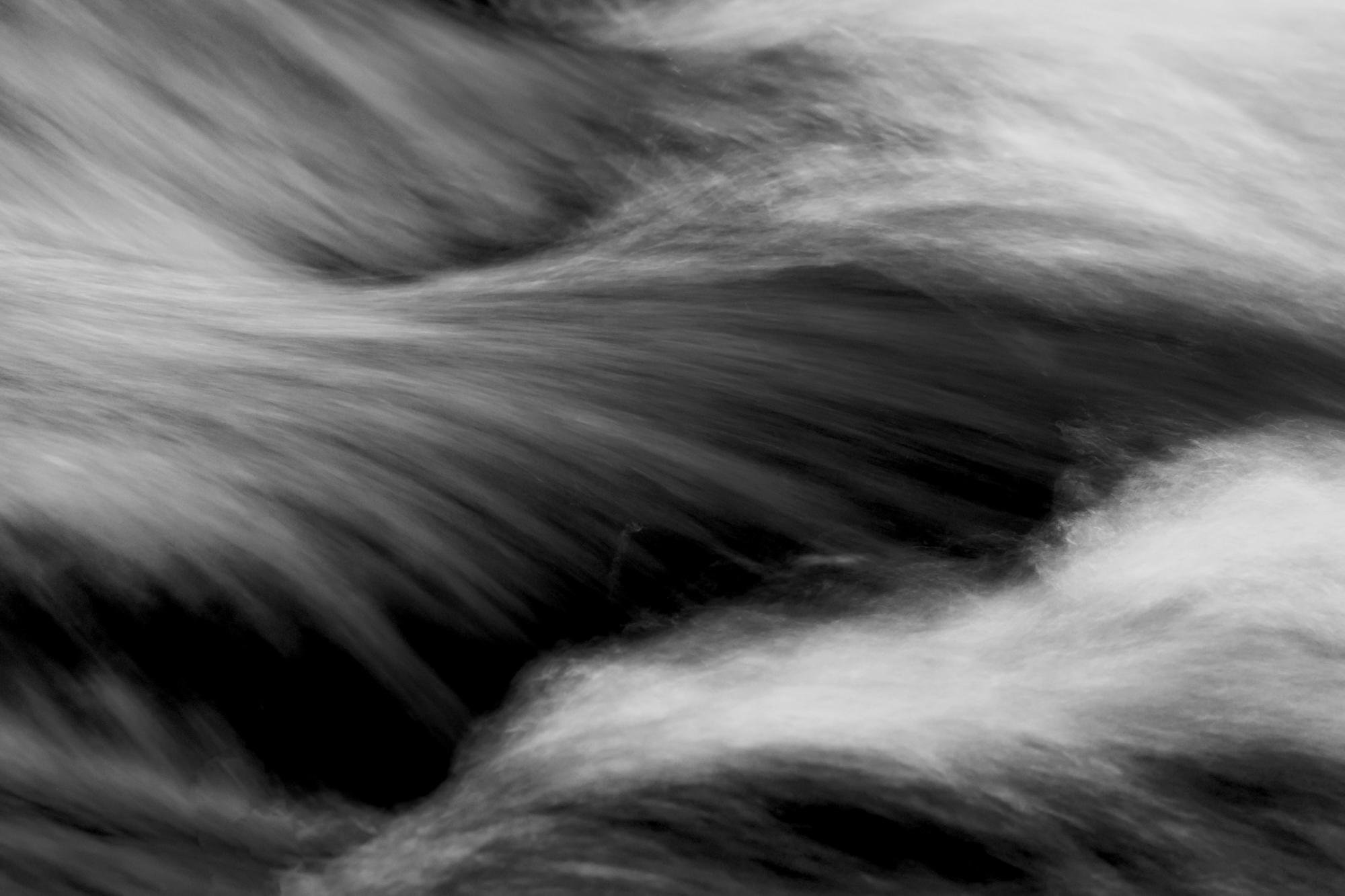 Long-exposure black-and-white close-up of flowing water streaking diagonally across dark rocks, creating blurred, silky bands and shadowed channels.