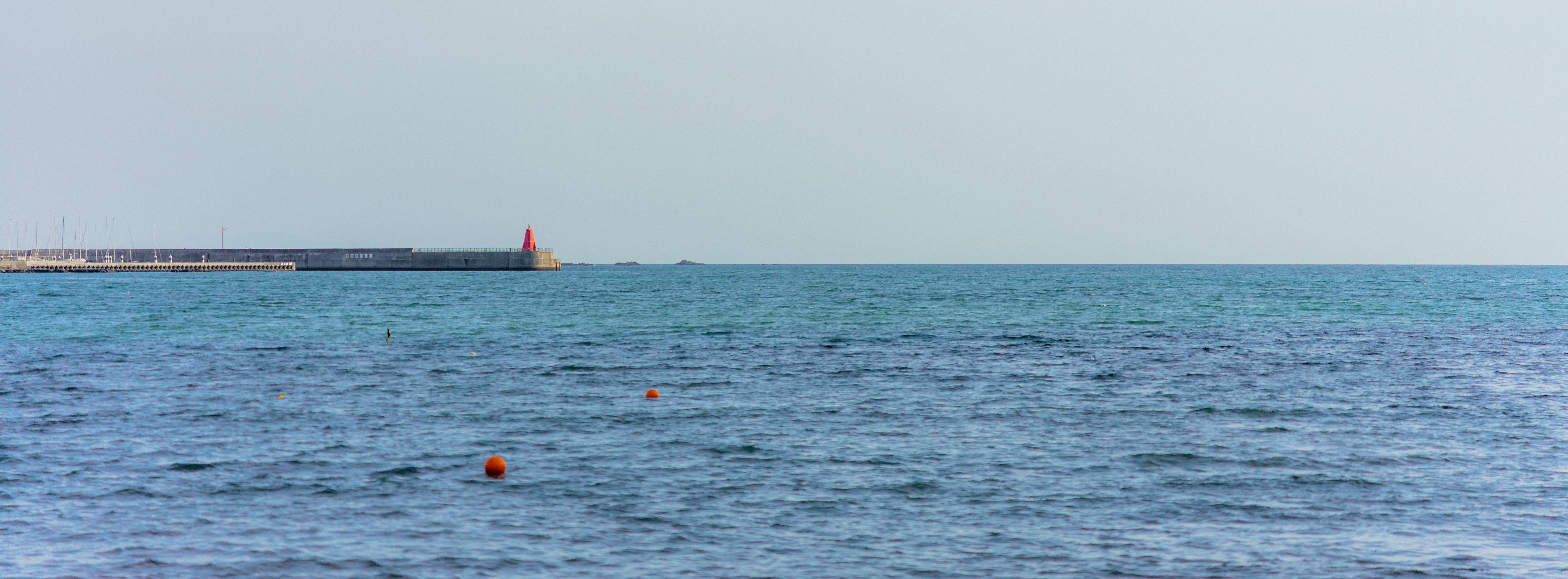 The image shows a calm sea with a few scattered orange buoys on the water. In the background, there's a breakwater with a red structure, possibly a lighthouse or marker, on top. The sky is clear and blends softly with the horizon.