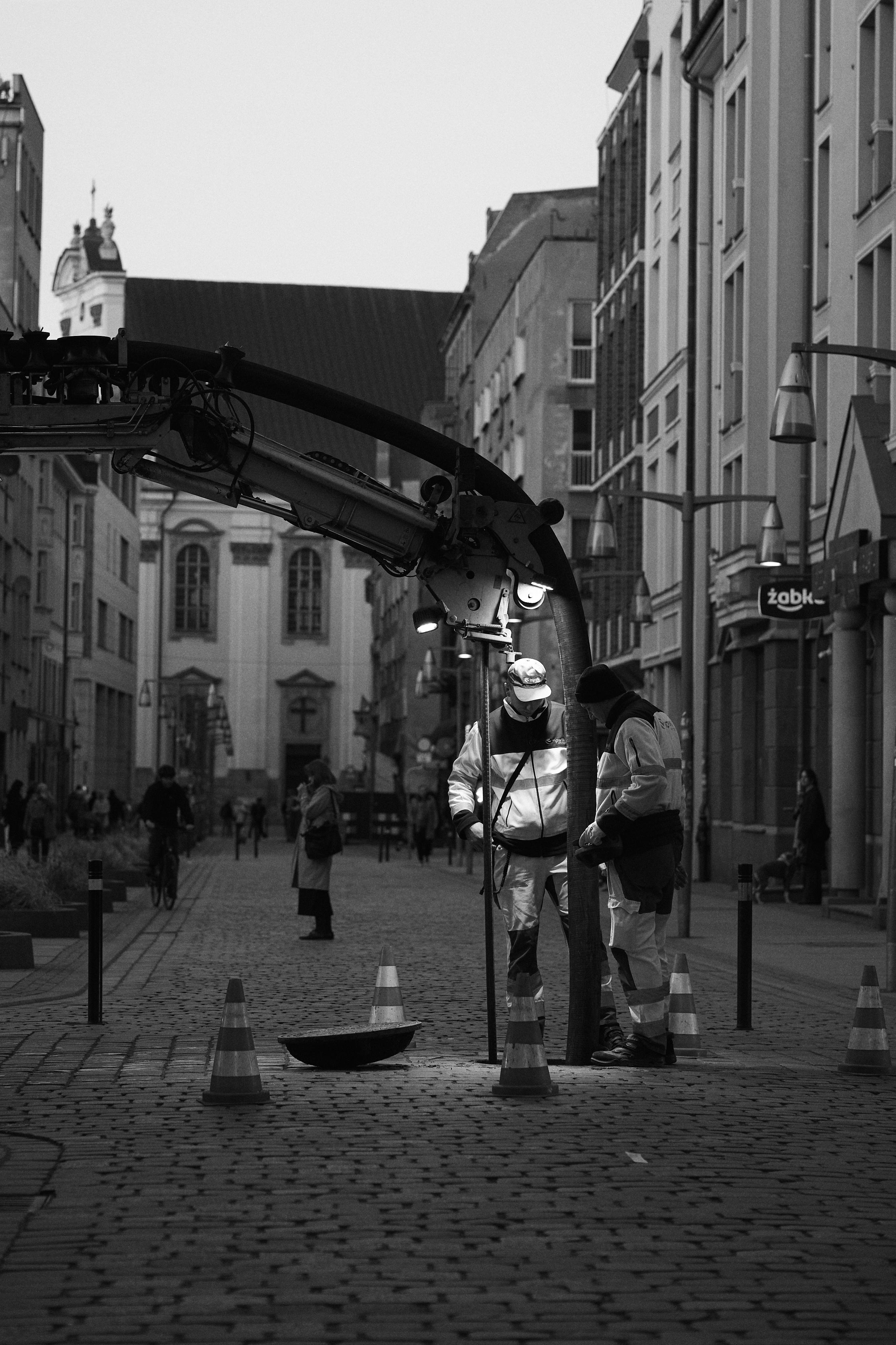 Two road workers in reflective jackets stand on a cobblestone street beside a large vacuum excavator arm and hose, with traffic cones marking a work area; pedestrians, cyclists, and multi-story buildings line the street in the background.