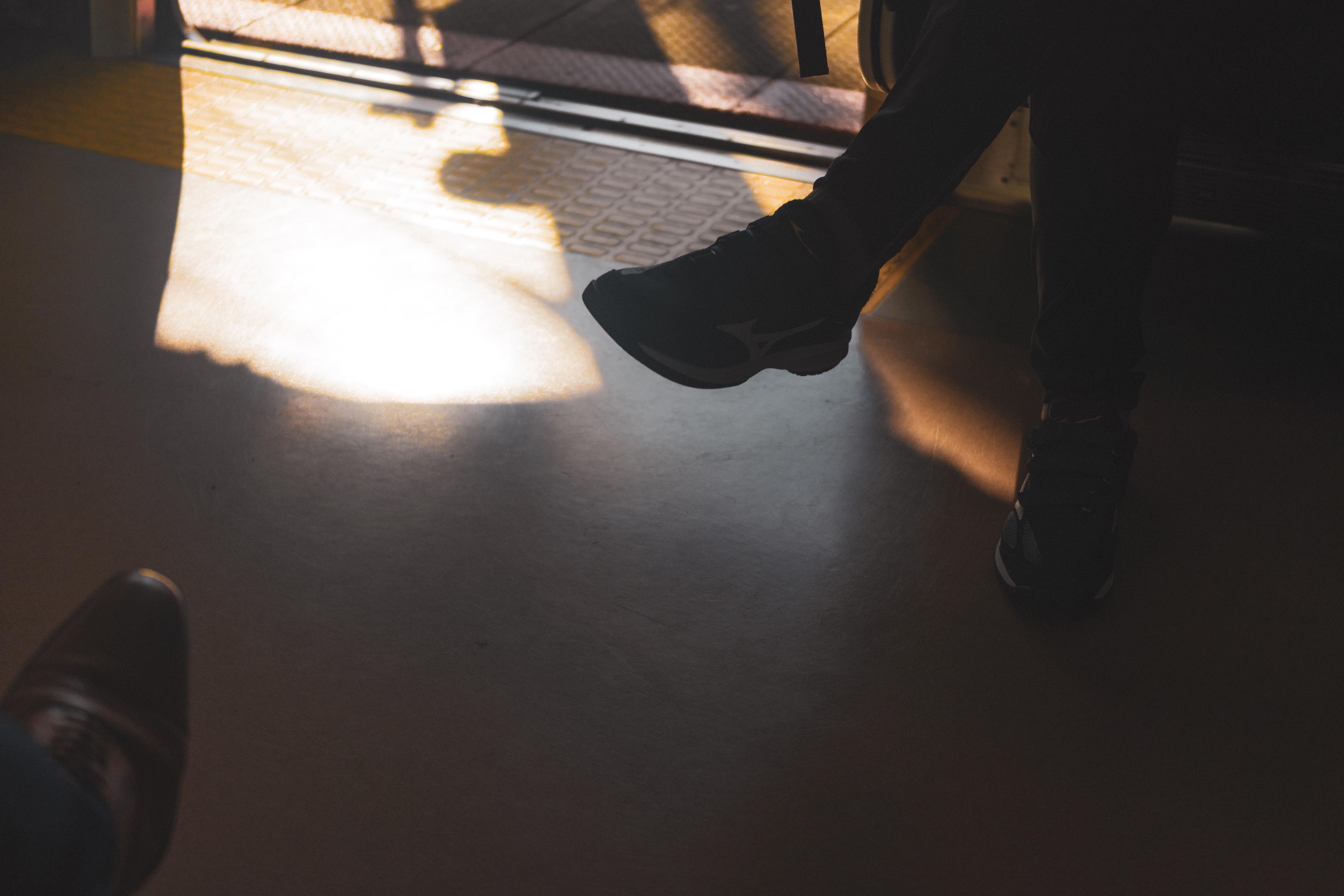 The image shows a dimly lit interior scene, possibly in a vehicle or train, with partial views of two people. One person is sitting with their legs crossed, wearing dark pants and sneakers, while the shadows and sunlight create patterns on the floor. The setting suggests a moment of travel or commute.