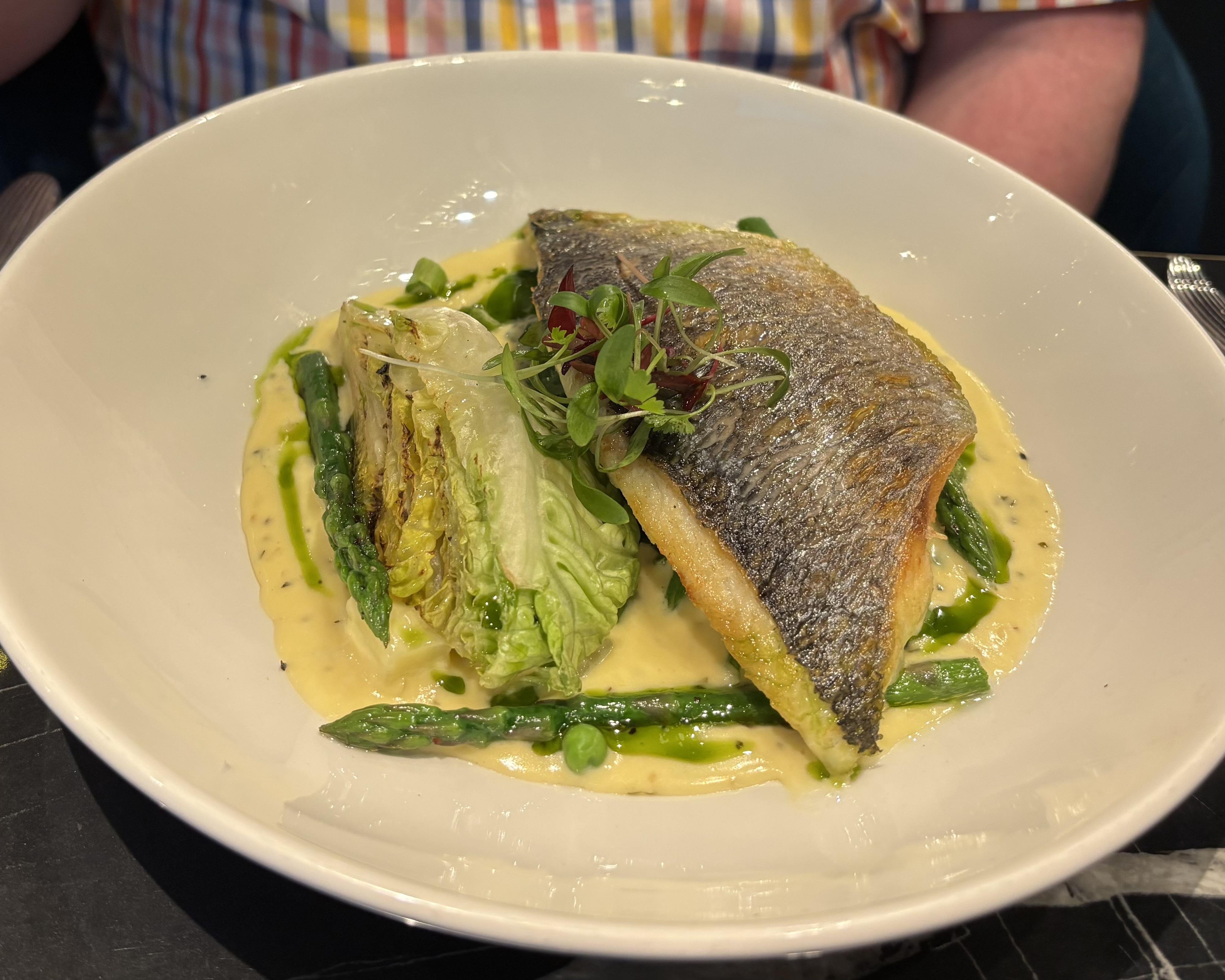 The image shows a dish featuring a fillet of fish, likely seabass or similar, atop a bed of creamy sauce. Accompanying the fish are asparagus spears and a wedge of grilled or roasted lettuce. The dish is garnished with microgreens, adding a touch of color and freshness.