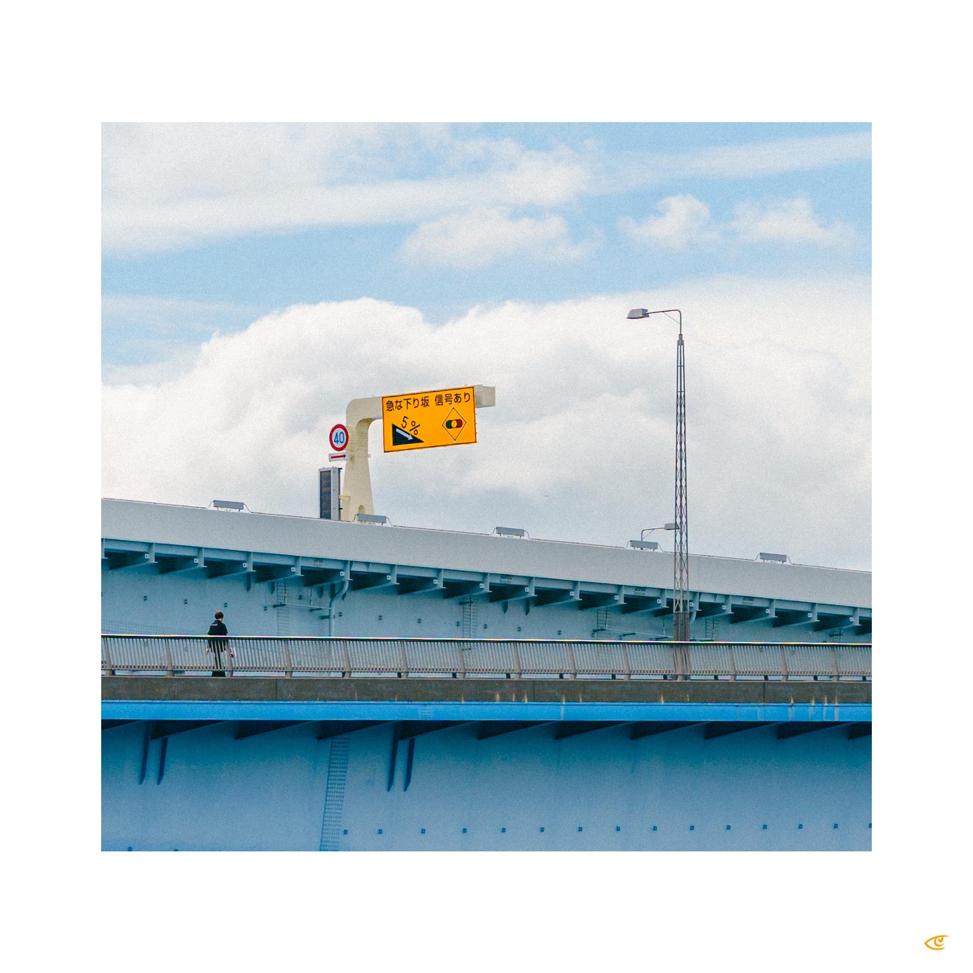 Pedestrian walkway on a large blue bridge with a lone person walking along the railing; overhead yellow road sign with Japanese text and a 40 speed limit sign against a cloudy sky.