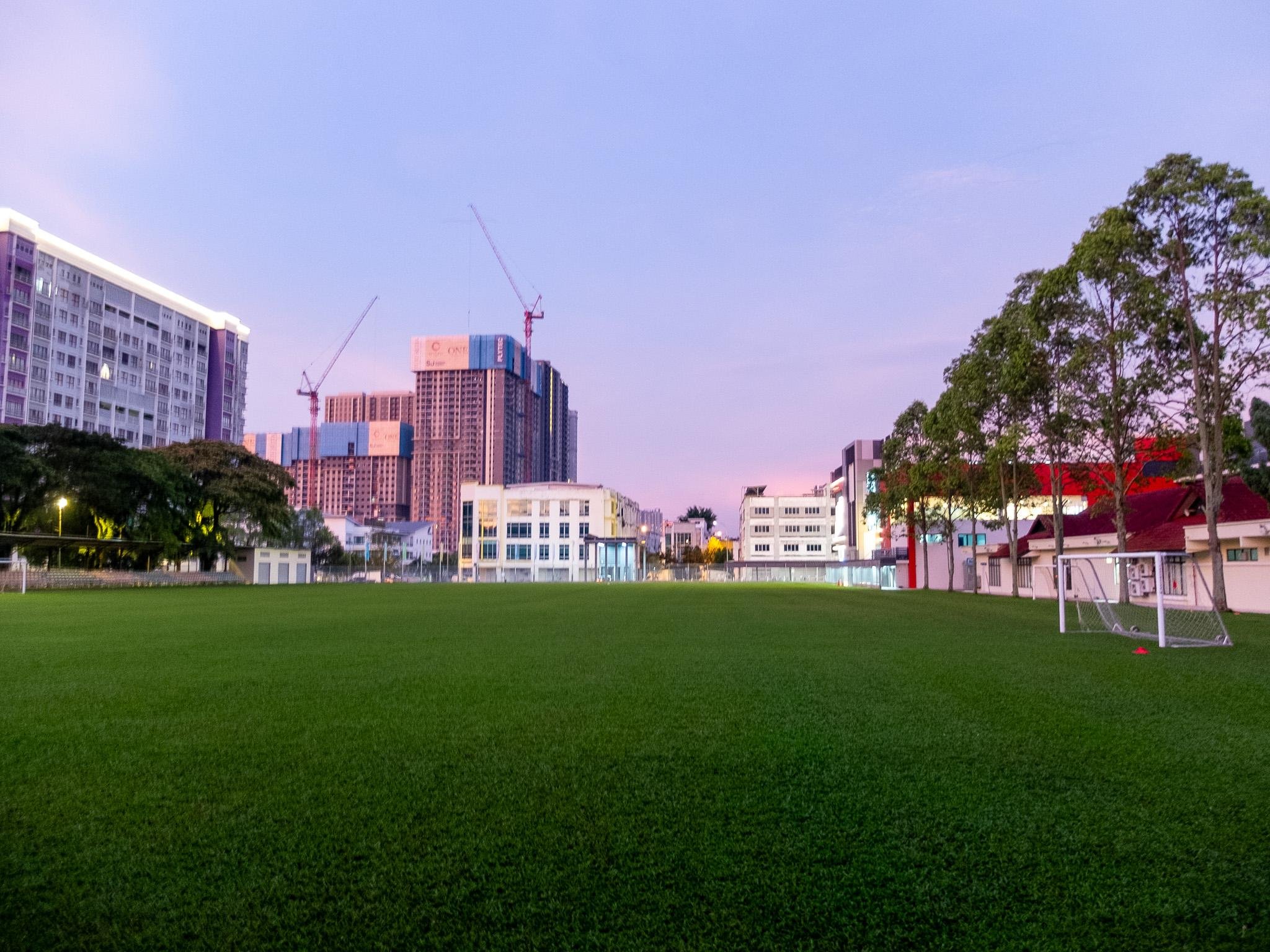 The image shows a large grassy field, likely a sports field or park, with a goalpost visible on the right side. In the background, there are several buildings, some under construction with cranes visible. The scene is set during twilight or early morning, as evidenced by the soft lighting in the sky. Trees line the right side of the field.