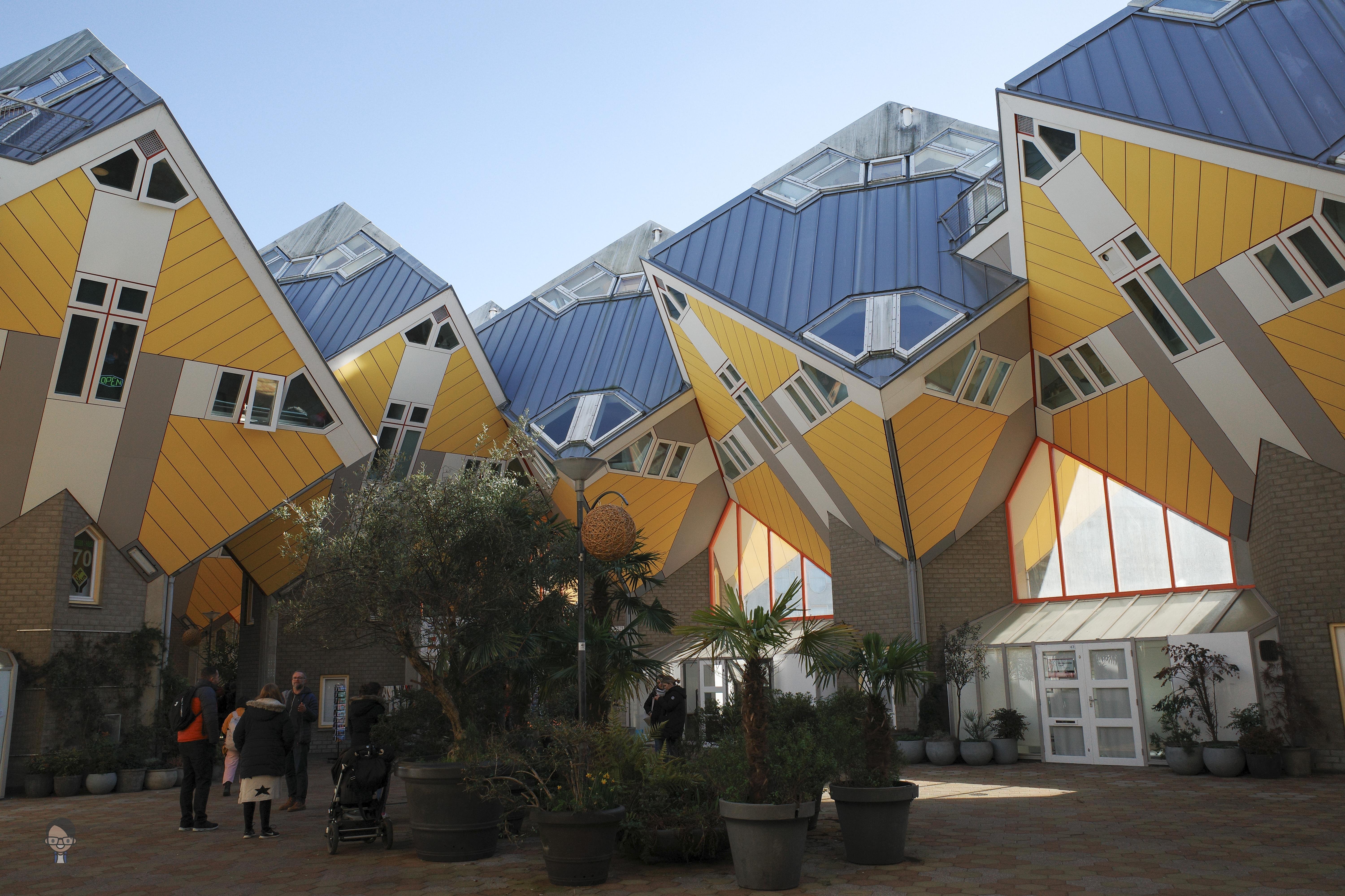 The image shows a group of uniquely designed cube-shaped houses. These are known as the Cube Houses in Rotterdam, Netherlands. Designed by architect Piet Blom, they are tilted at 45 degrees and set upon hexagon-shaped pylons, creating a visually striking architectural effect. The buildings have bright yellow and blue facades and are surrounded by people and plants in the foreground.
