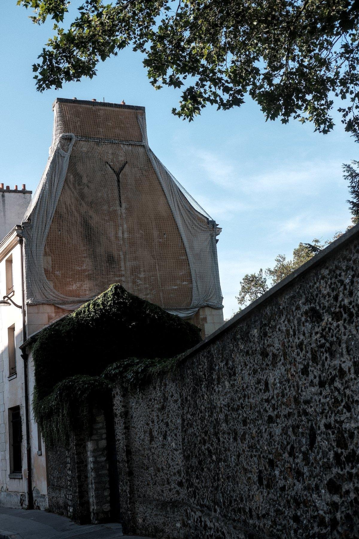 Photo verticale. Dans une rue, vue sur un côté d’un petit immeuble, en brique rouge, avec des cheminées. Il y a un filet qui le recouvre, et dessous, une toile blanche tombant à gauche et à droite. Un espèce de Y est en haut au centre, et le bas de la toile dessine un arrondi sur le bas, de chaque côté. Ce Y et la toile, avec la couleur rougeatre fait penser à un énorme décolleté. Au premier plan, un mur en pierre allant vers cet immeuble
