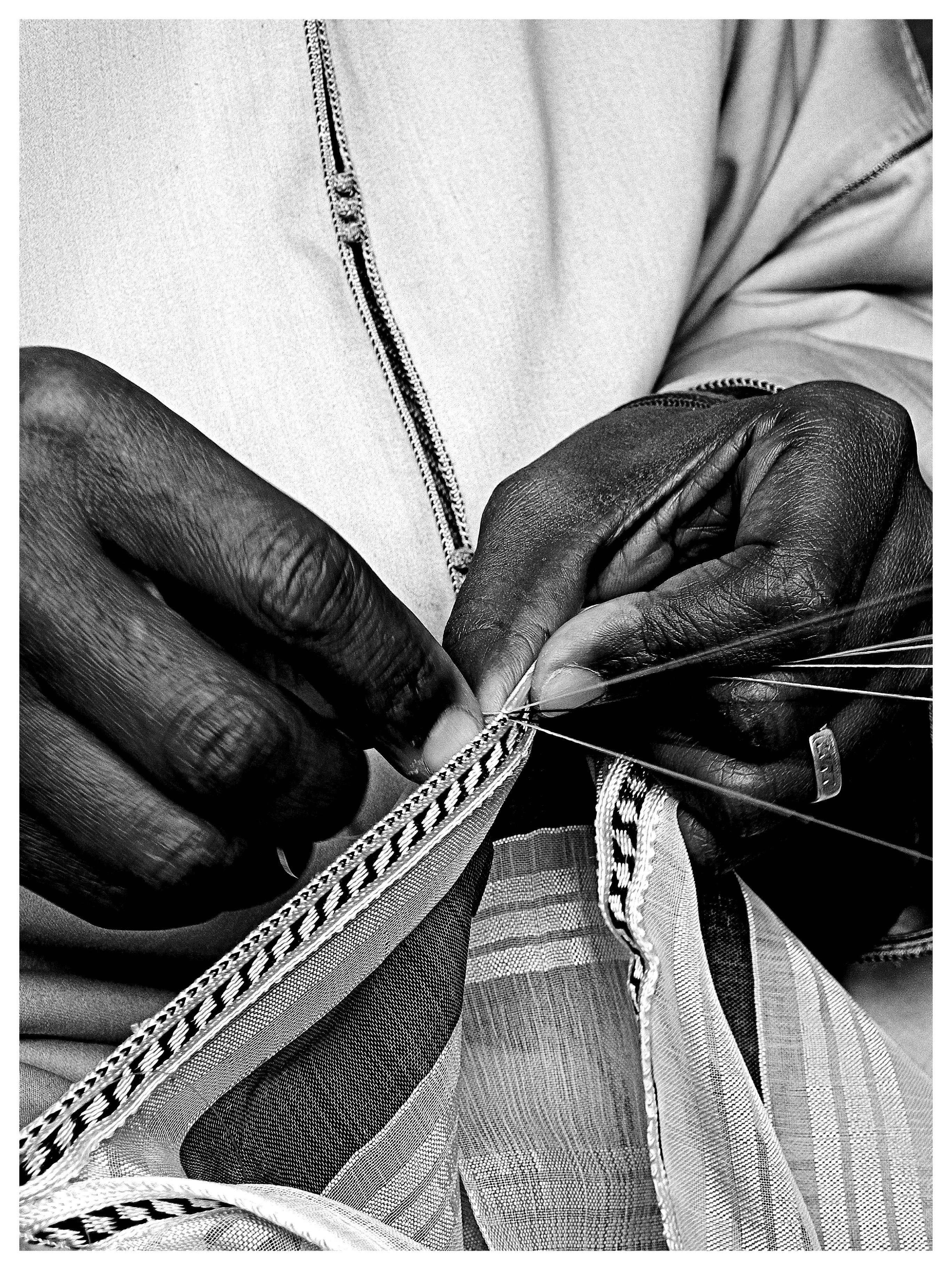 Close-up black-and-white photo of two hands sewing patterned trim onto striped fabric with a needle and thread; part of a zippered garment visible in the background.