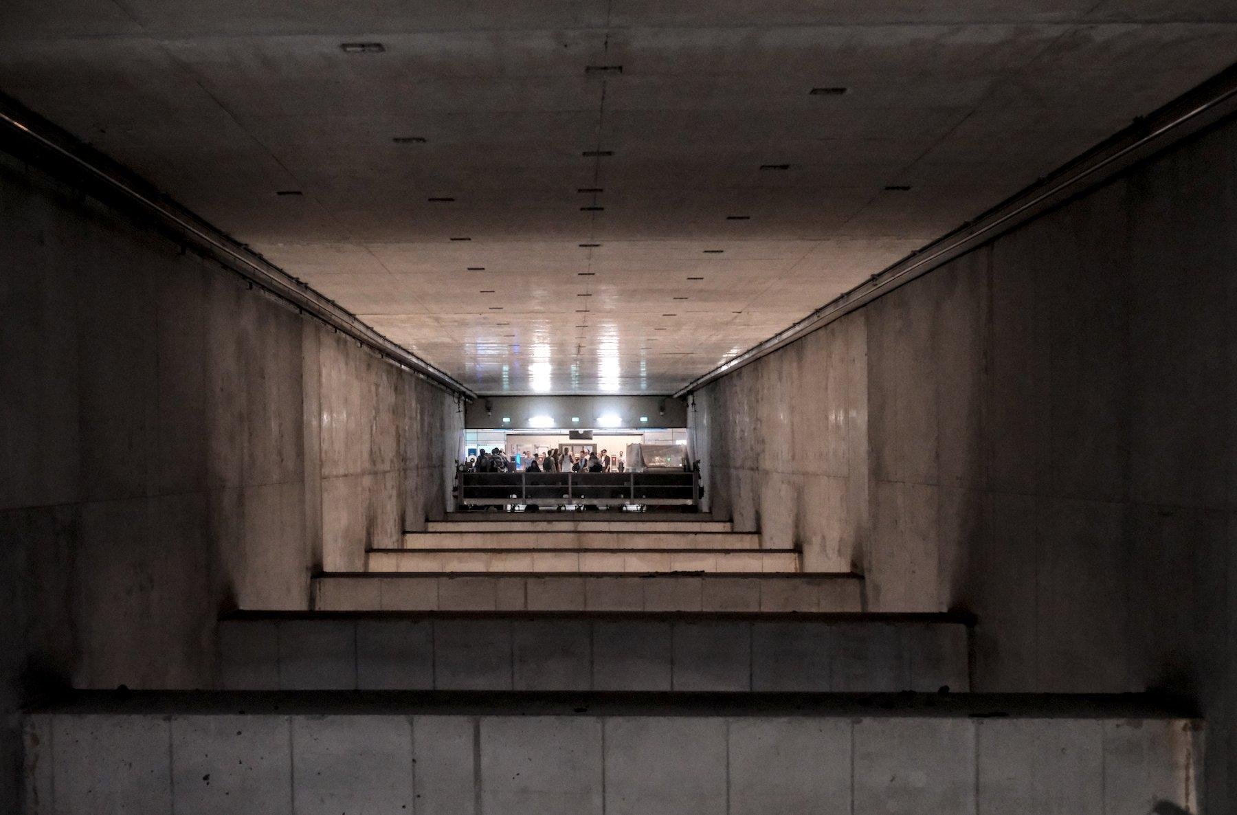 Photo horizontale. Vue sur une autre entrée du métro en face d’un grand vide avec des poutres horizontales traversant le vide à interval régulier dans un long passage en béton