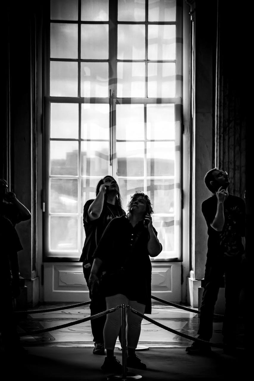 Silhouetted group of people inside a building, standing behind a rope barrier in front of a tall multi-pane window; several look upward, with the bright outdoor light backlighting the scene.