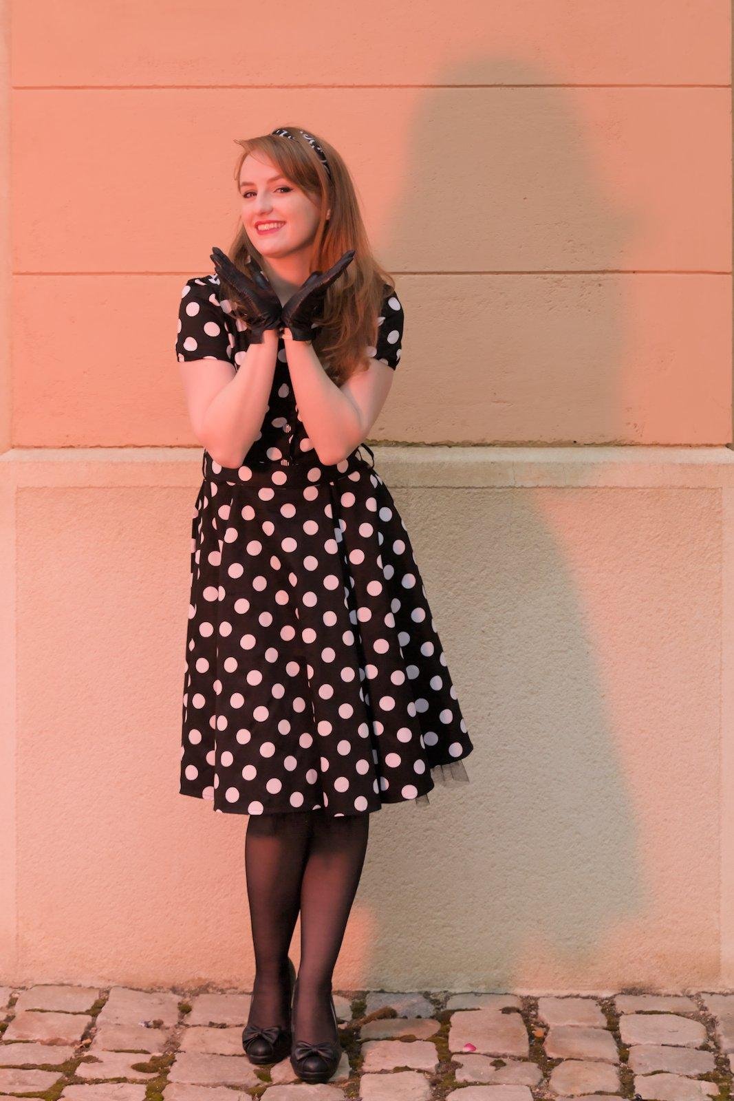 Une jeune femme aux cheveux long roux, posant devant un mur blanc, portant une robe noire à pois blanc, des collants noirs, des chaussures noires avec un petit nœud, des gants noirs et un serre tête noir avec des motifs blancs. Ses mains forment un V sous son menton et sa tête est tournée un peu vers sa droite, les yeux vers la caméra et un sourire enjoué