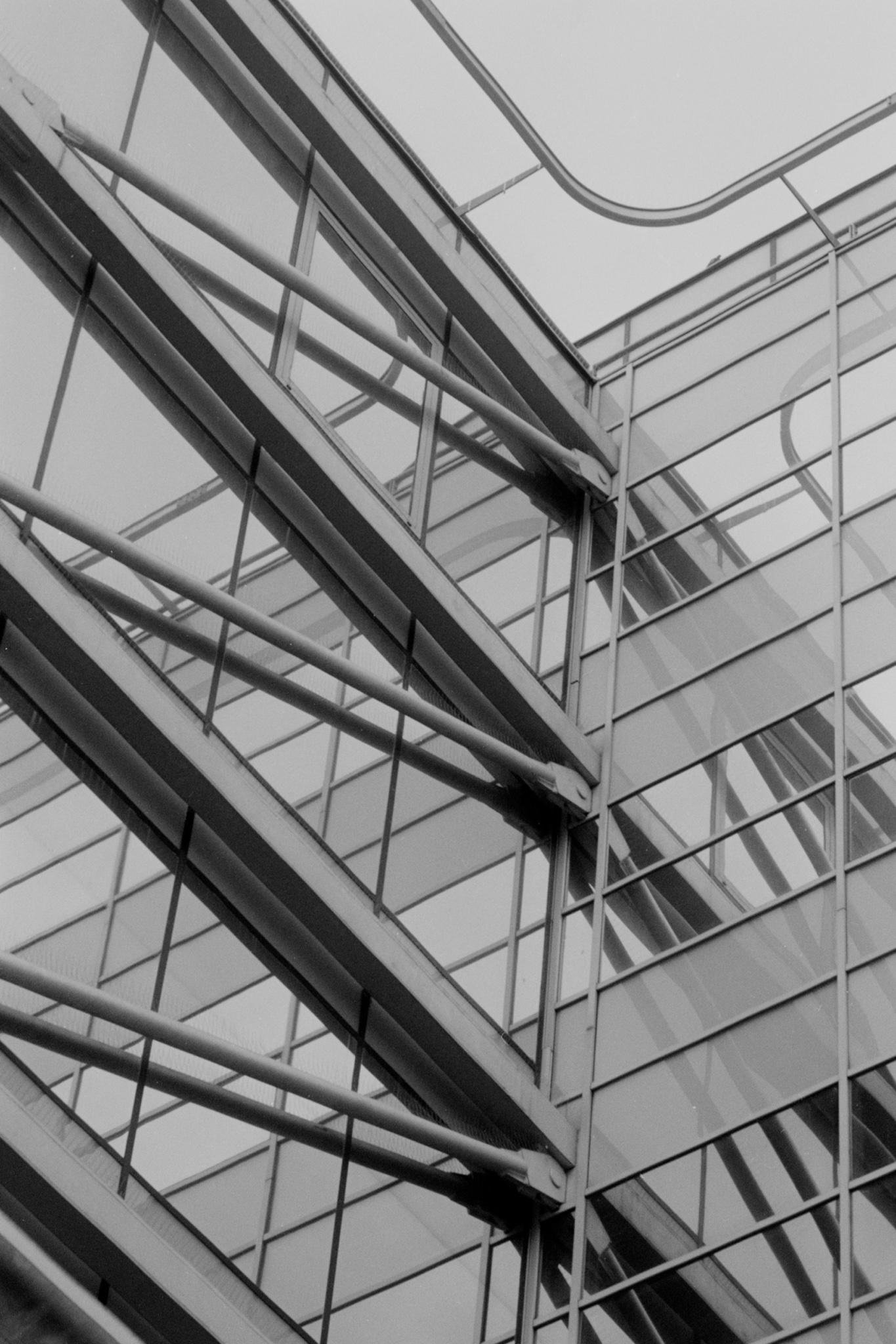 Upward view of a modern glass-and-steel building facade with diagonal beams, grid-like window panels, and curved metal framing near the roofline in black and white.