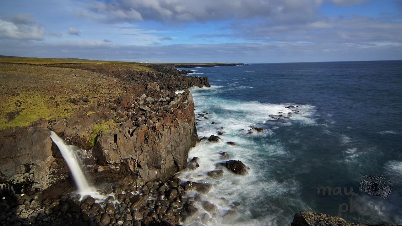 Rocky coastal cliff with a small waterfall dropping onto rounded black stones, waves breaking against scattered rocks along the shoreline, and a wide ocean stretching to the horizon under a cloudy sky.