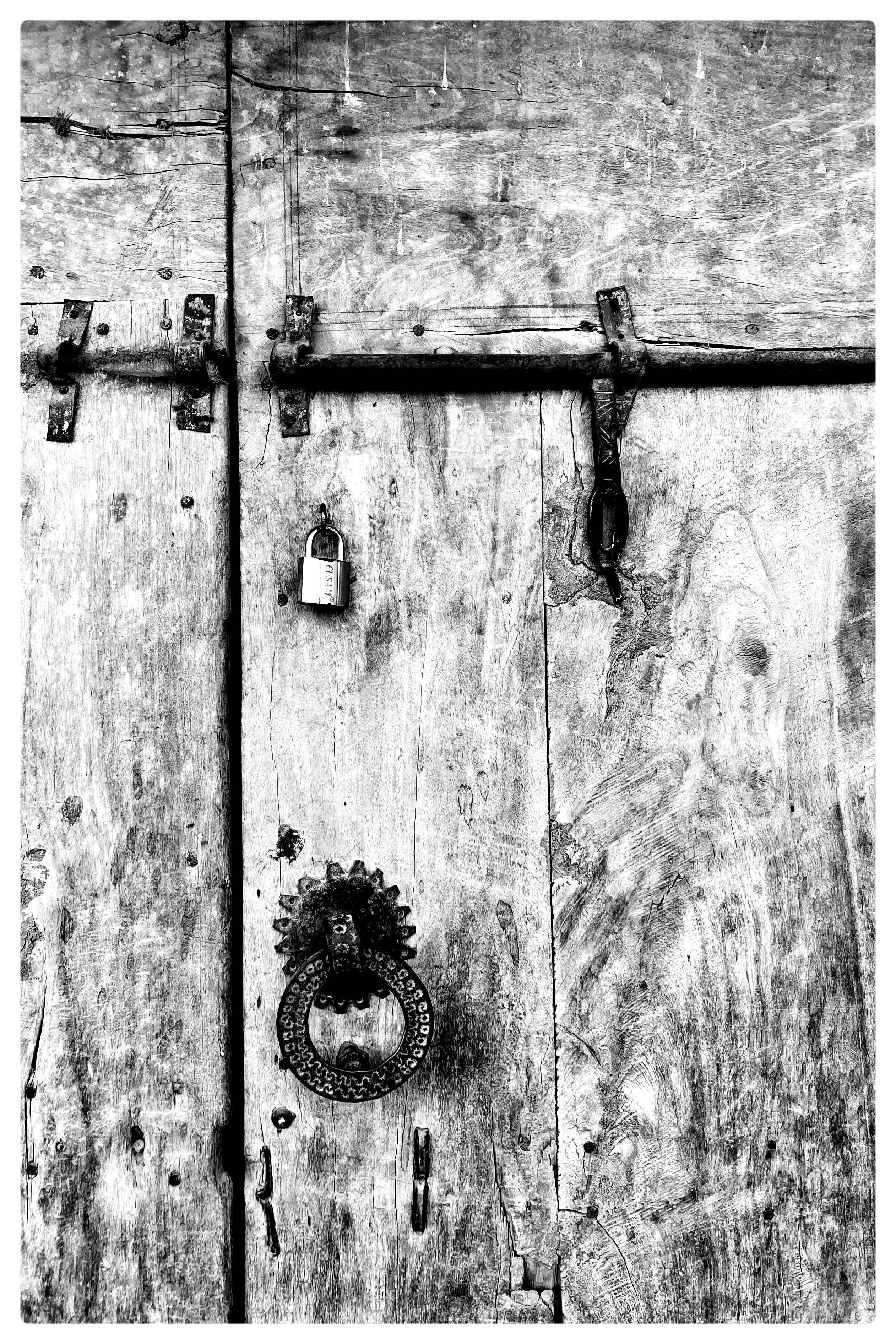 Weathered wooden door with visible grain and texture, secured by a metal latch and padlock at the top. A circular, ornate metal door knocker is attached below. The door's surface shows signs of aging and wear. Black and white photograph.