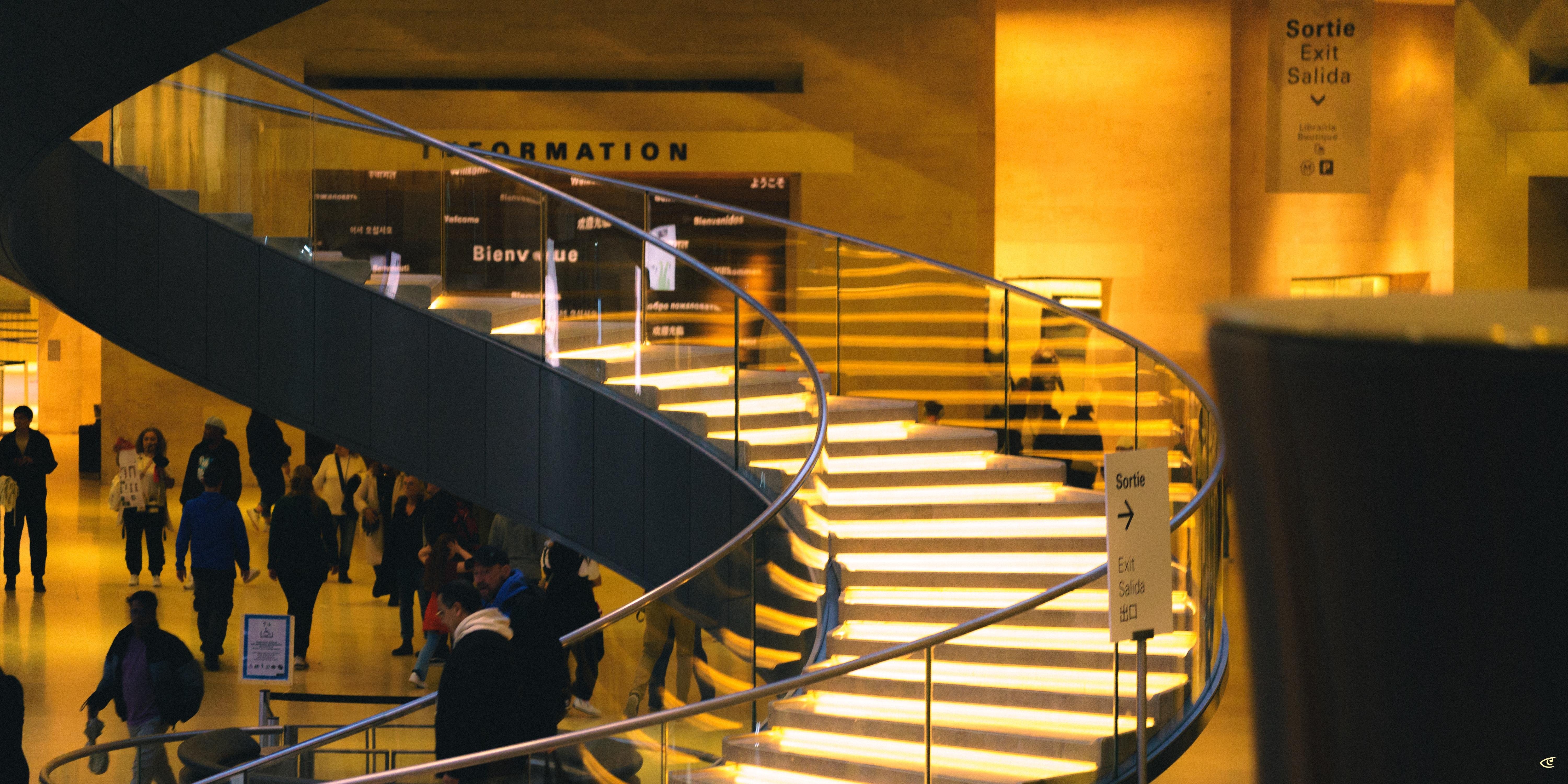 Curved, illuminated staircase with metal railings inside a large building, with people walking on the floor below and signs reading “Information” and “Sortie / Exit / Salida” in the background.