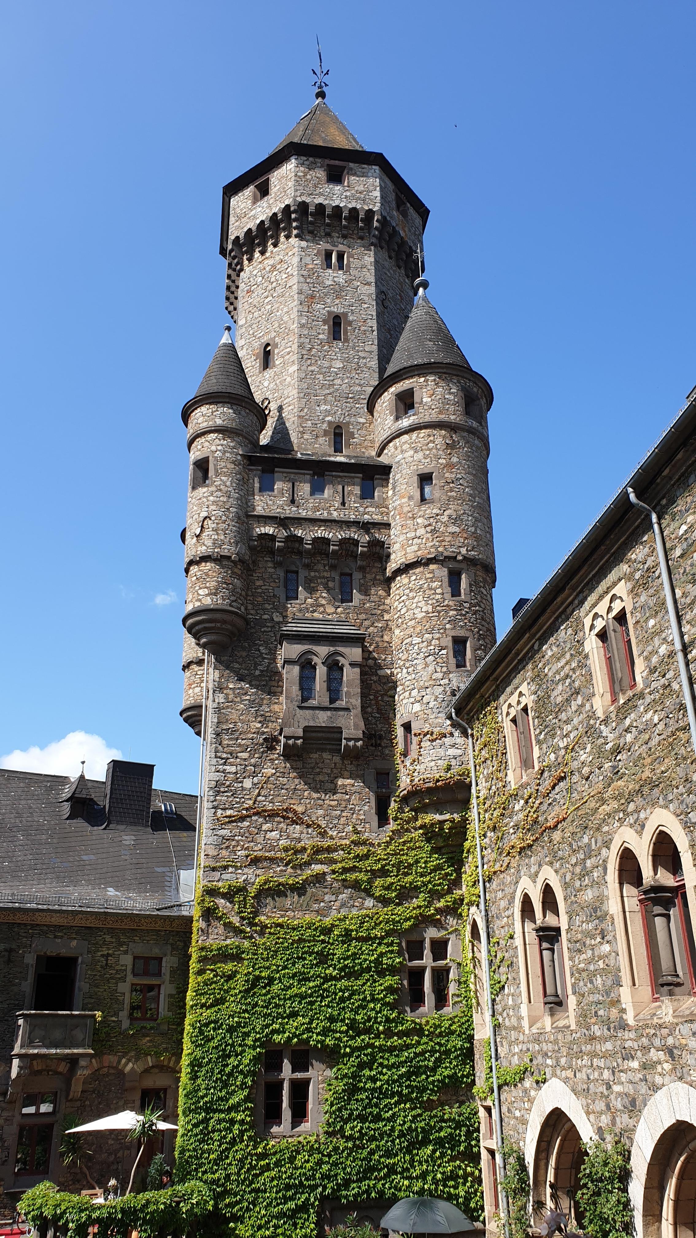 Photo taken at Schloß Braunfels, showing a striking upward view of the castle’s main keep framed by bright blue sky. The tall, cylindrical stone tower dominates the scene, built from rough grey-brown masonry and featuring narrow slit windows and an octagonal roof crowned with a weather vane. Attached to the central tower are several round turrets with pointed conical roofs, giving the structure a fairy-tale appearance. This keep, one of the castle’s most iconic parts, dates back to medieval fortifications and was later expanded during Gothic and Baroque renovations, serving both defensive and residential purposes. Below, the castle walls are covered in lush green ivy, with arched windows and reddish stone frames visible among the foliage. To the left and right, additional wings of the castle enclose a sunny courtyard where a parasol and plants hint at a small terrace café.