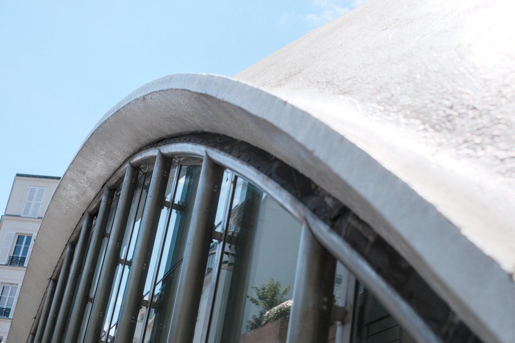 Photo horizontale. Gros plan sur une arche de béton blanc qui a l’air de débuter un dome vers la droite hors cadre. Dessous est une baie vitrée protégée par des barreaux en acier. Dans le coin supérieur gauche, le ciel bleu