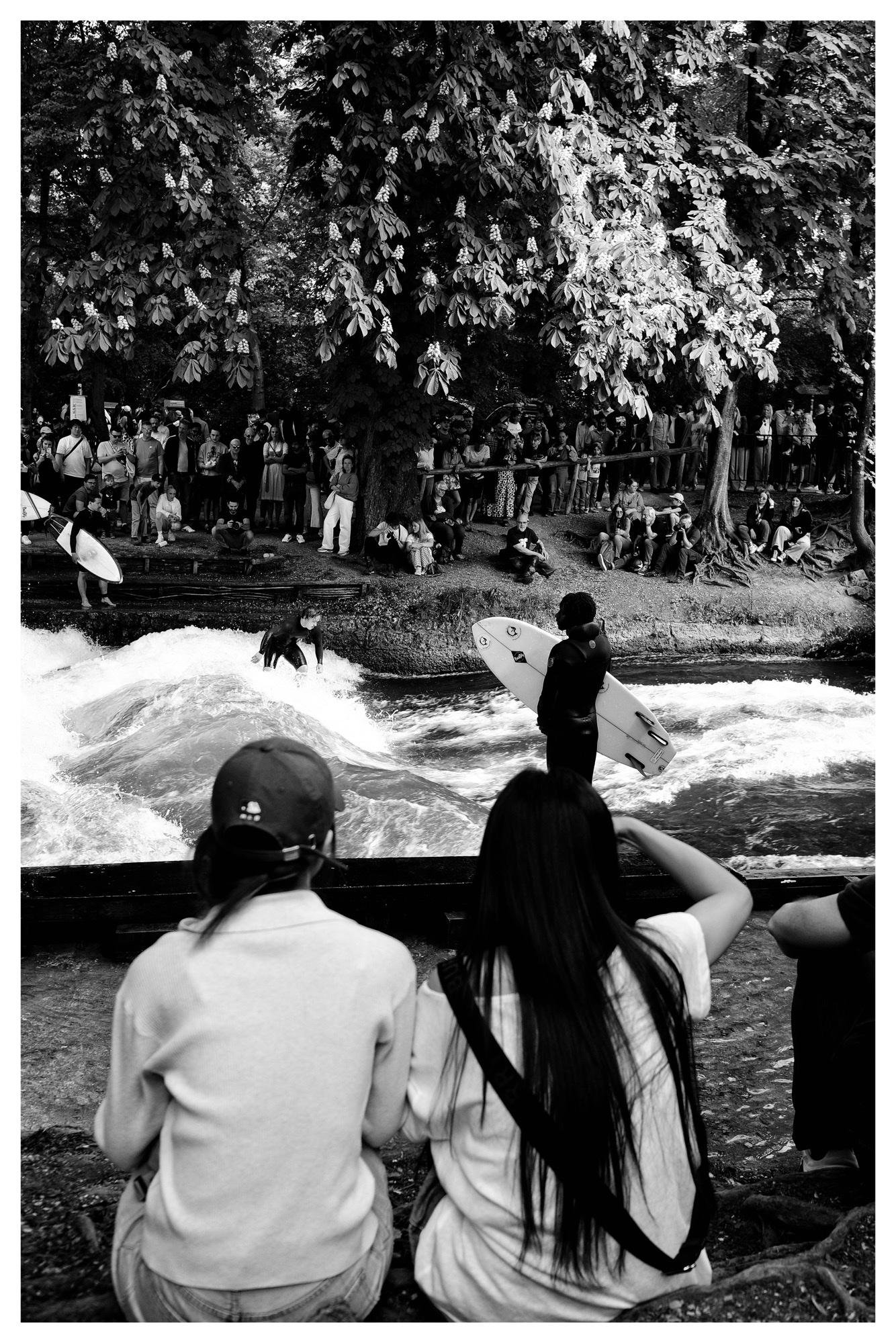 Black-and-white photo of a river surfing spot: two seated spectators in the foreground, a standing surfer holding a board at the water’s edge, another surfer riding a standing wave, and a large crowd watching from the tree-lined bank behind.