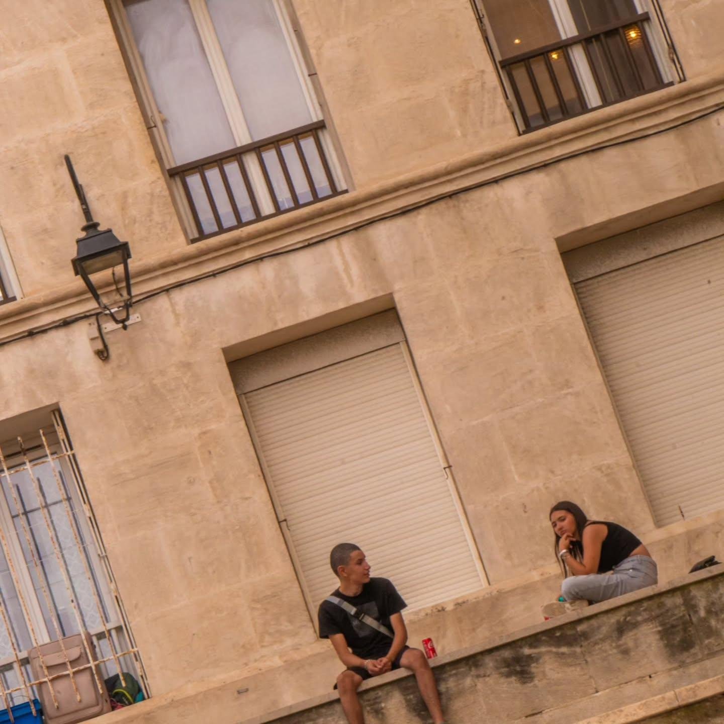 L'image montre deux personnes assises sur un rebord devant un bâtiment aux murs beiges. Le bâtiment dispose de plusieurs fenêtres et d'un lampadaire. La personne de gauche porte un t-shirt et un short noir et est assise de front, la tête tournée à droite vers l'autre personne. L'autre est dans un haut noir avec un jean gris, assise en tailleur sur le rebord, vers la gauche, mais regarde en trois quarts. Il y a une canette de soda à côté de celle de gauche. La perspective de l'image est inclinée.