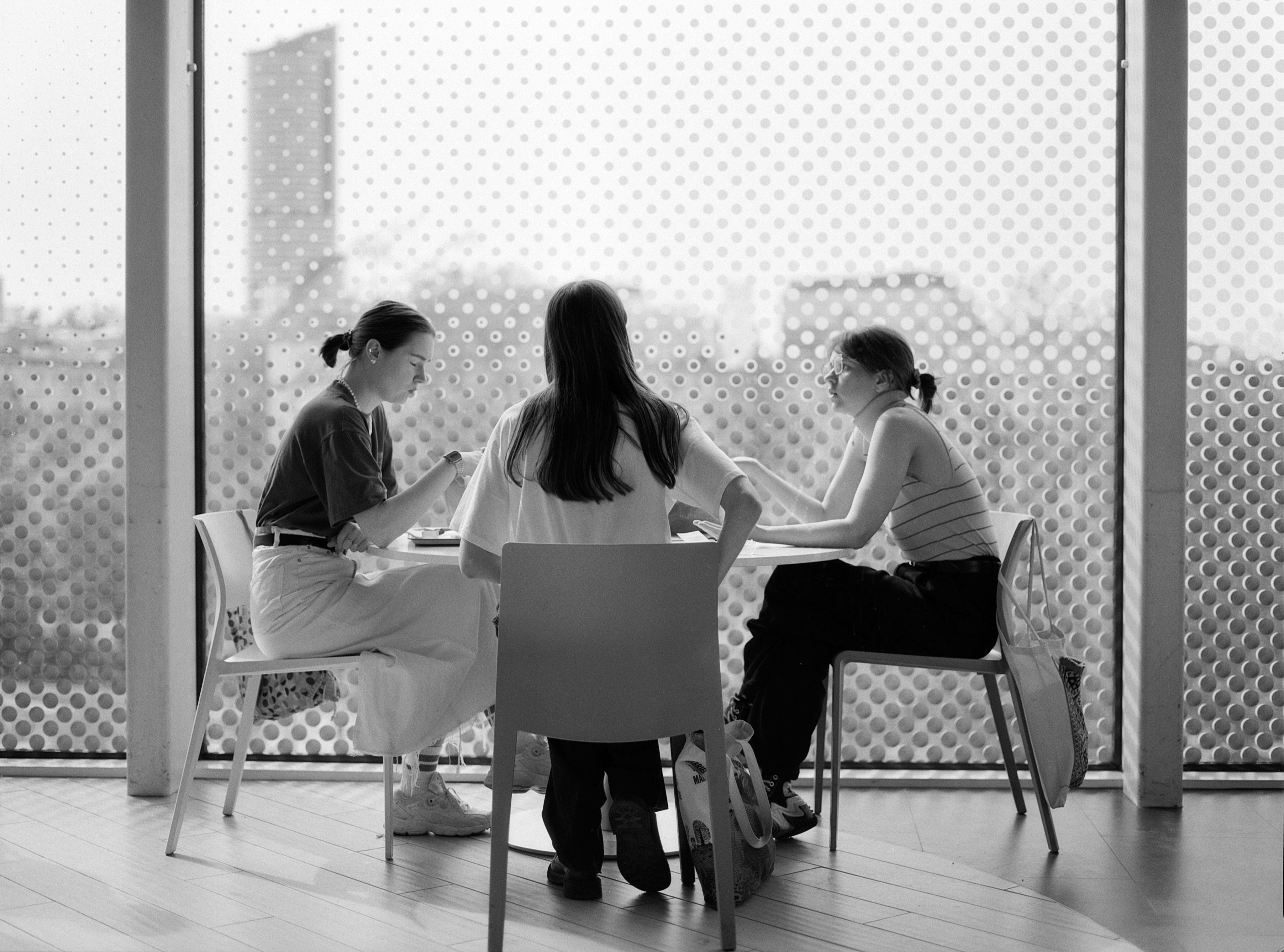 Three people sit at a table, engaged in discussion. One person is wearing a long skirt and has their hair tied back, another has long hair and a T-shirt, while the third wears a striped top and glasses. Bags rest on the floor beside them. The window behind provides a blurred view of a cityscape. The image is in black and white.