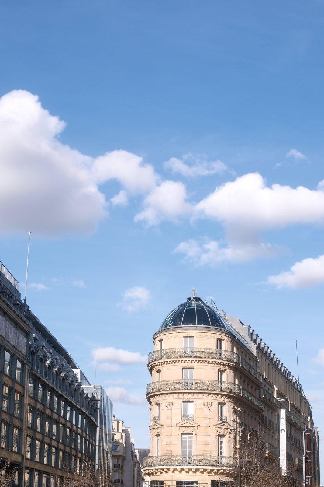 Photo verticale. Deux immeubles parisien, celui de droite est étroit, arborant une coupole en verre à son sommet, sous un ciel bleu avec quelques nuages