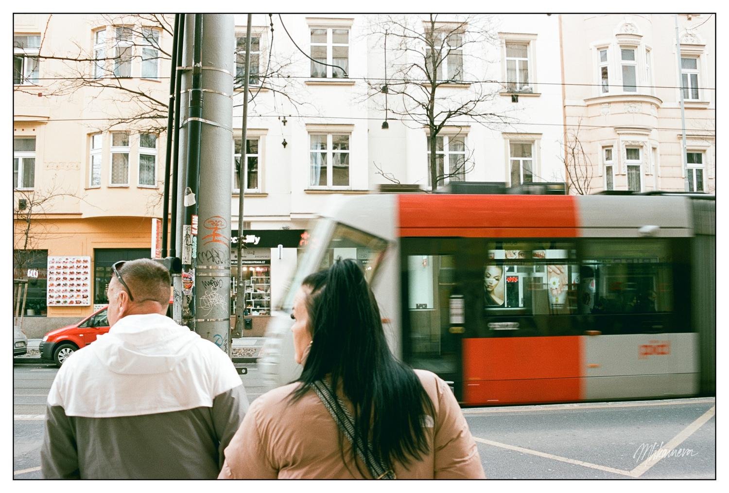 Two people stand on a sidewalk facing away, observing a red and white tram moving across the street. The background features multi-story buildings with pale facades and several windows. A street light pole with graffiti is visible next to the people. The scene captures a moment in an urban setting, with bare trees and a parked car in the distance.