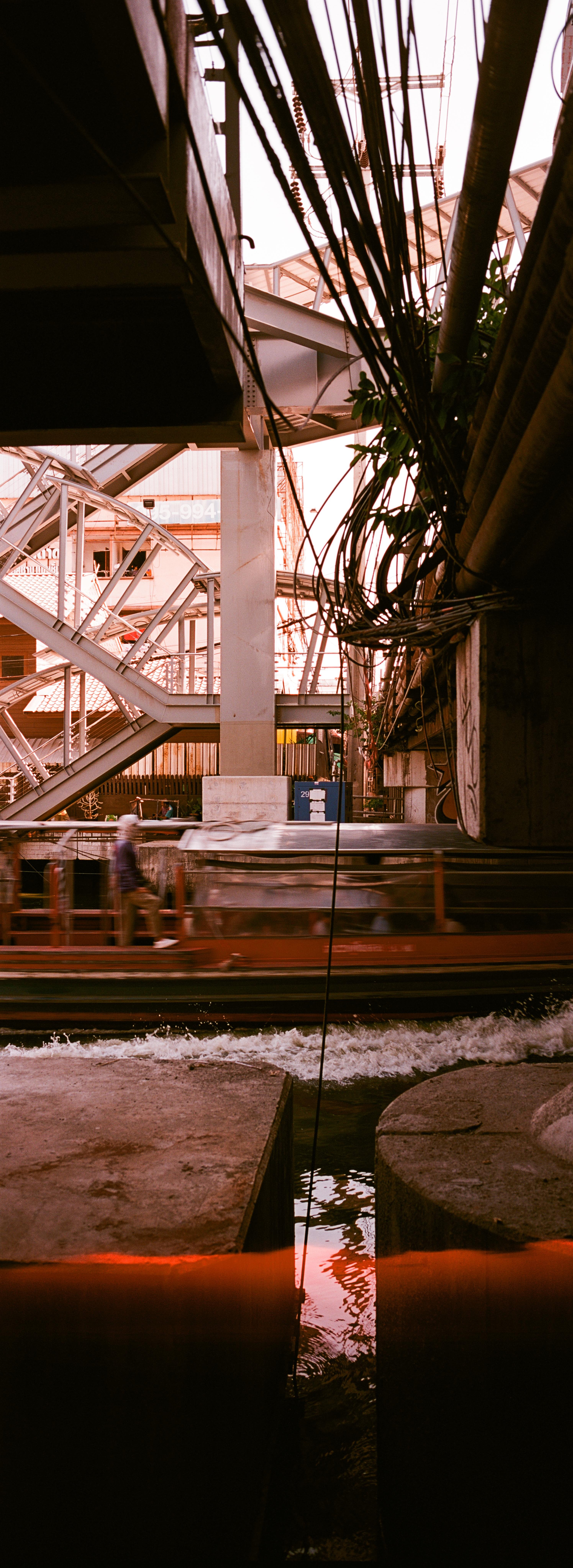 EN: vertical panoramic photo in color with very strong red tints of a blurred boat passing in front of concrete installation and under a tangle of electrical wire. On the other side, a complex staircase structure rises above the canal
FR: photo panoramique format portrait, en couleur avec des rouges très marqués, dd'un bateau flou qui passe devant des structures en béton et sous un enchevêtrement de fils électriques. De l'autre côté s'élève une straucture assez complexe qui est en fait un escalier
