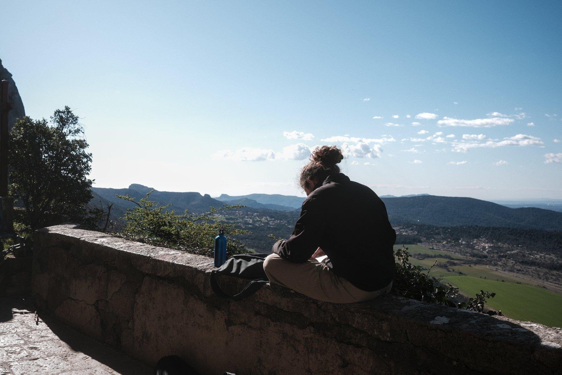 Photo horizontale. Un homme au cheveux long en chignon, assis en tailleur sur un parapet, un manteau et une gourde posés devant, tête baissée, probablement entrain de consulter son tél. En fond, un beau paysage aérien de la plaine au pied de la montagne