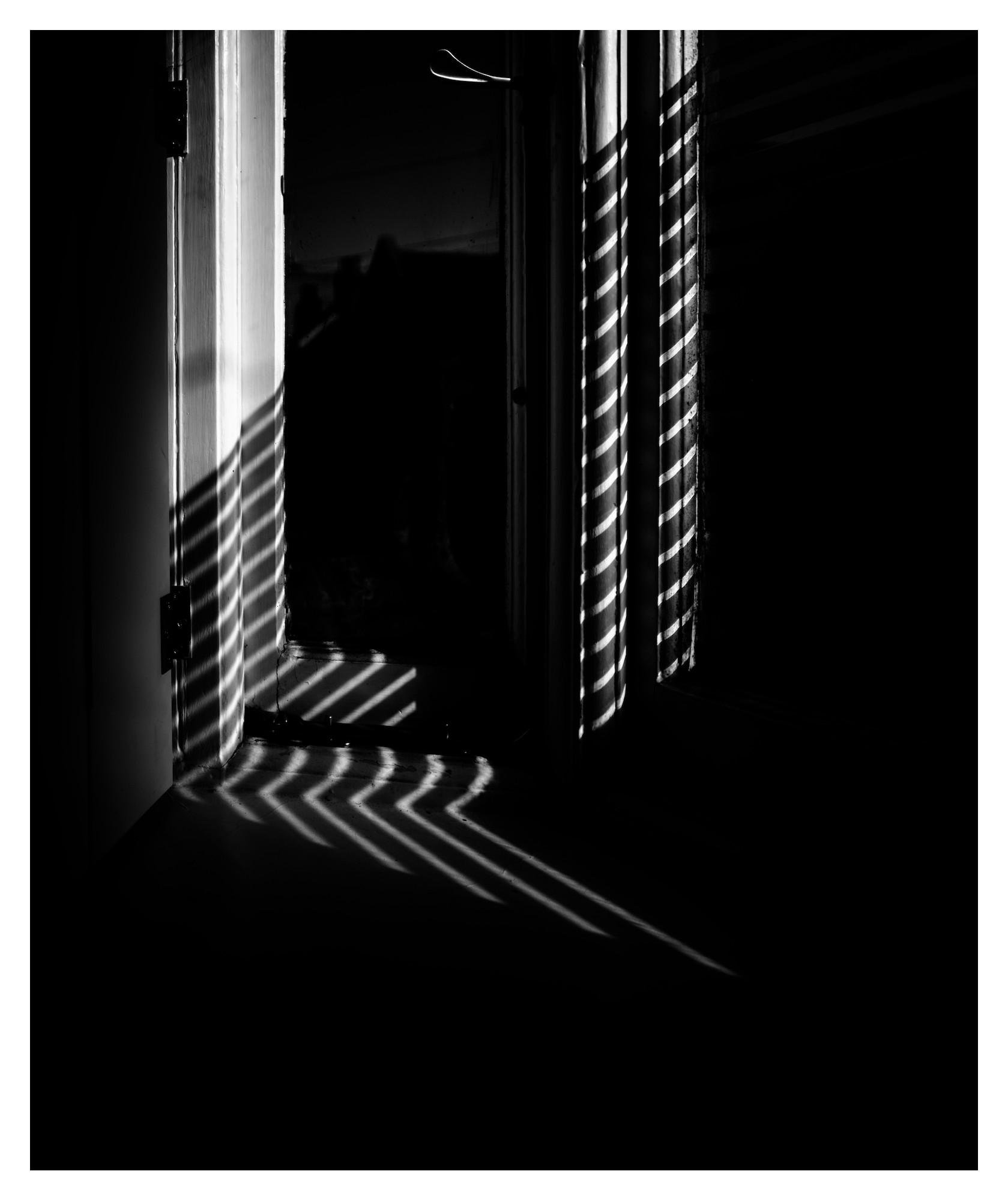 Black-and-white interior view of an open doorway and adjacent window, with bright diagonal stripes of sunlight from blinds cast across the doorframe and floor, leaving most of the room in shadow.