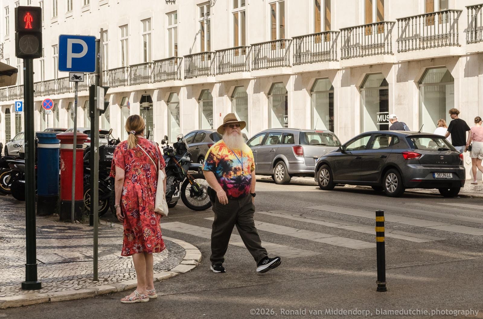 Two pedestrians at a city crosswalk: a woman in a red patterned dress standing near a parking sign and red pedestrian signal, and a bearded man in a tie-dye shirt and hat walking across. Parked cars and motorcycles line the street in front of a white building with many windows and balconies.