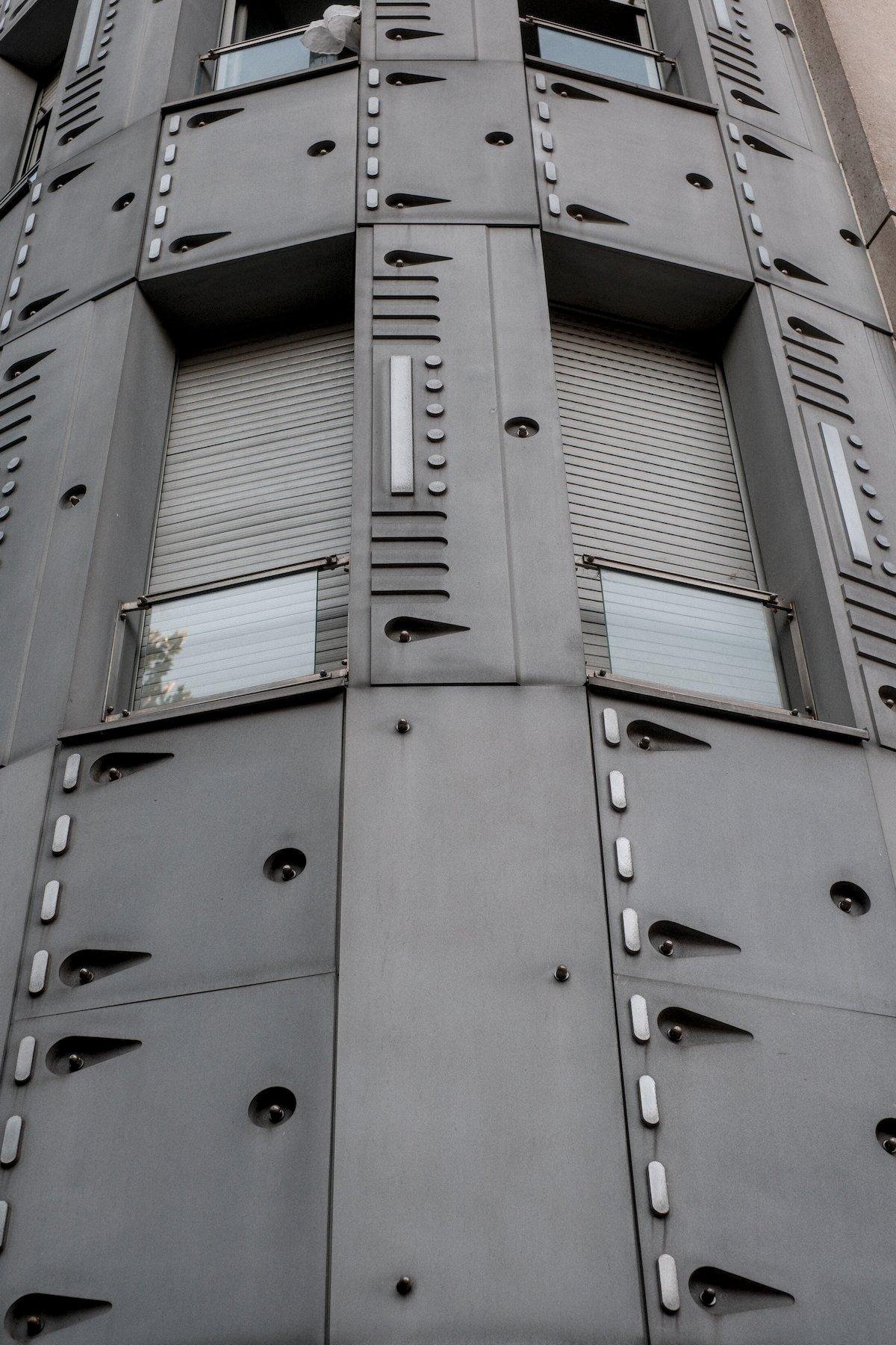 Photo verticale. Contre plongée sur des fenêtres étroites dans un mur courbé, recouvert de plaques grises faisant penser à du blindage