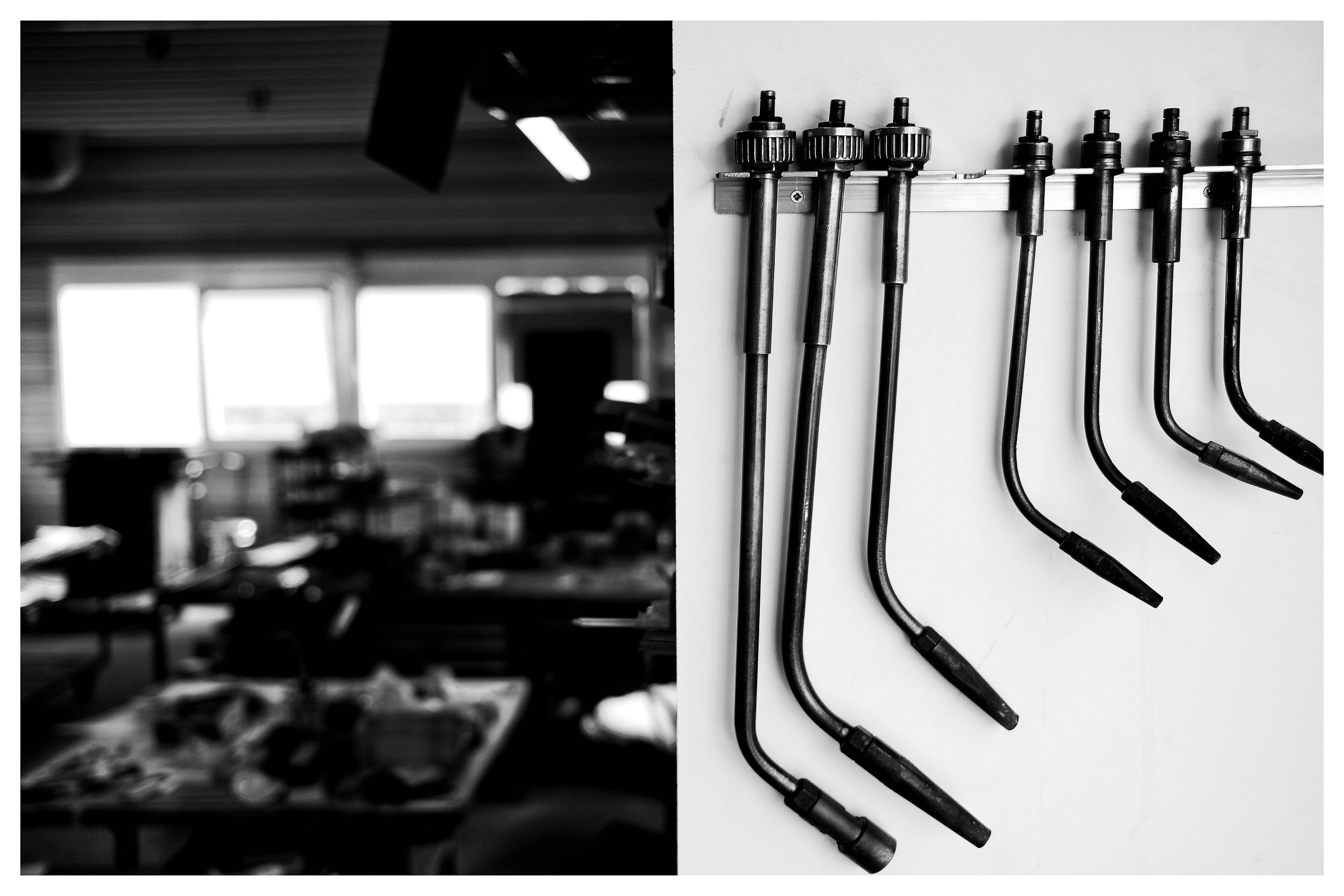 Black-and-white split image: blurred workshop interior on the left, and five metal welding torch handles with curved hoses hanging in a row on a white wall on the right.