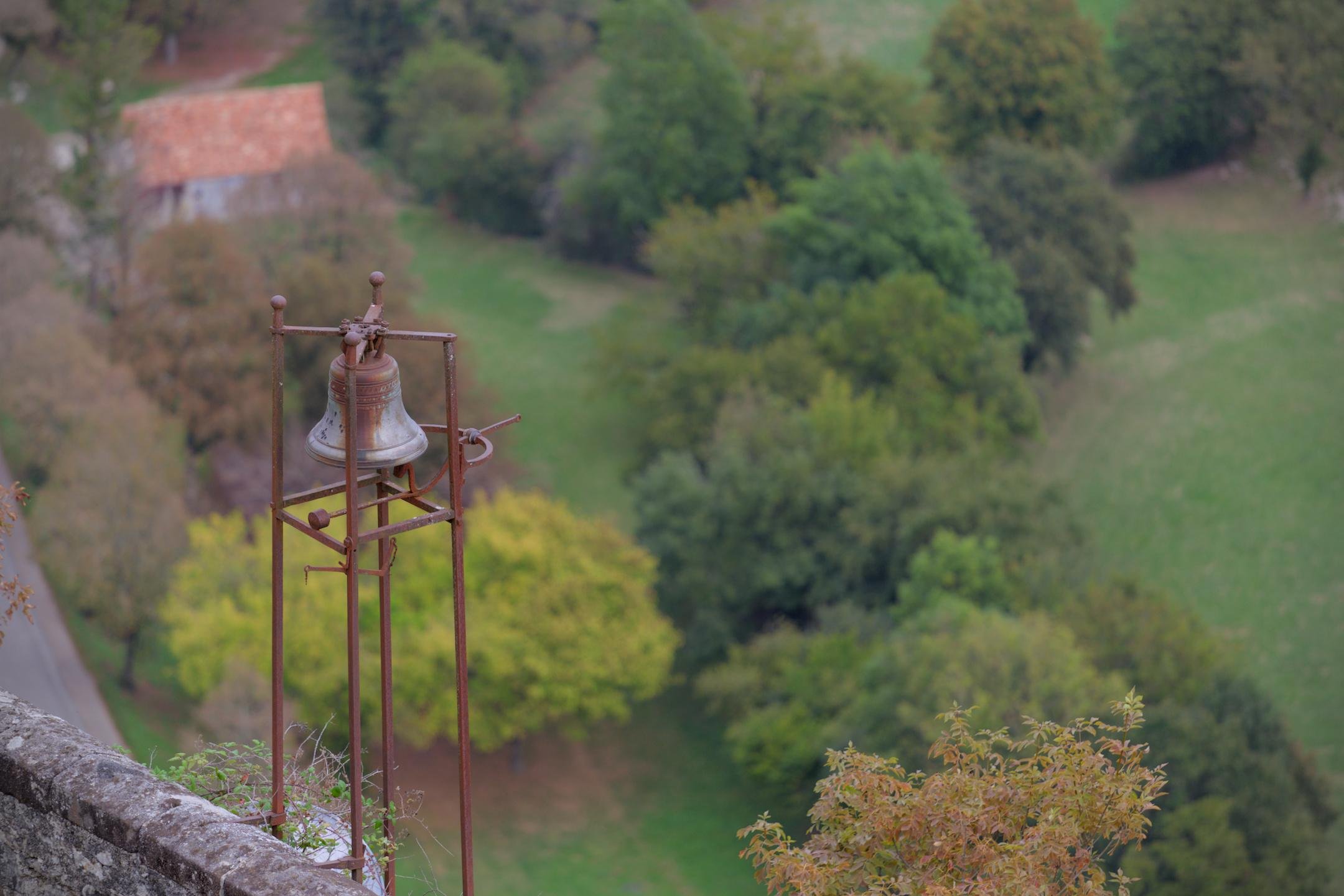 Photo format paysage montrant une vieille petite cloche dans son support rouillé en haut d’un mur, avec en contre bas, un paysage verdoyant