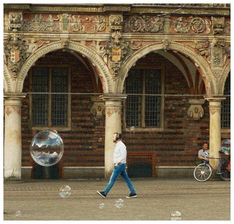 Man in a white shirt and blue jeans walking across a paved courtyard with soap bubbles floating nearby, in front of an ornate brick building with arched stone columns and tall windows; a bicycle and seated person are at the right edge.
