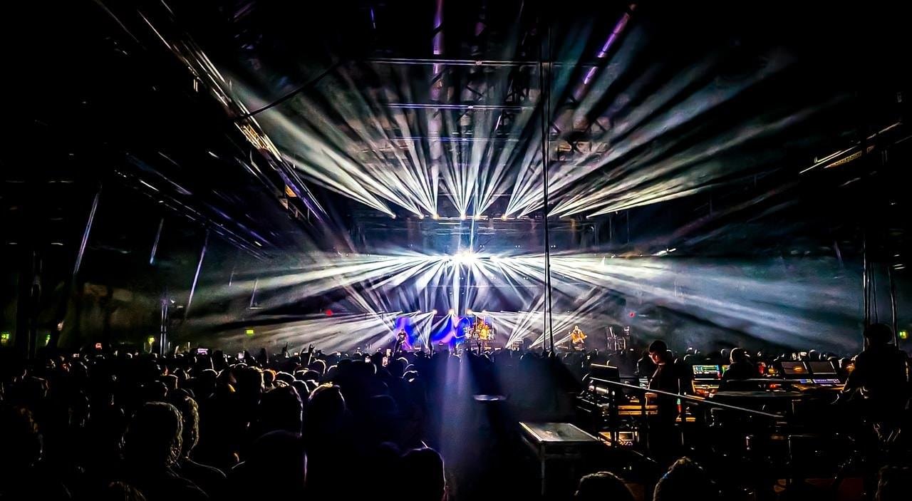 Wide shot of an indoor concert venue with a large crowd in silhouette facing a brightly lit stage; intense white beams radiate from overhead rigging, with performers and equipment visible on stage and a sound-mixing area on the right.